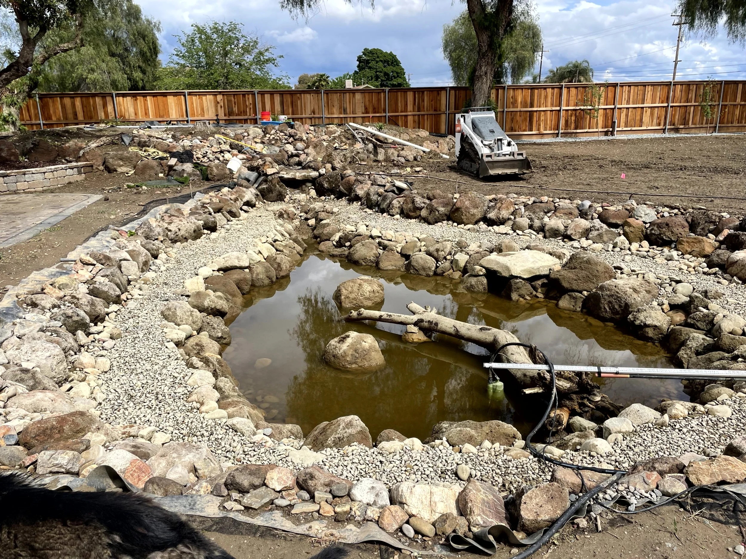 A backyard pond under construction, surrounded by rocks and gravel, with a small pond skimmer and construction equipment nearby, a wooden fence in the background, and some trees and cloudy sky overhead.