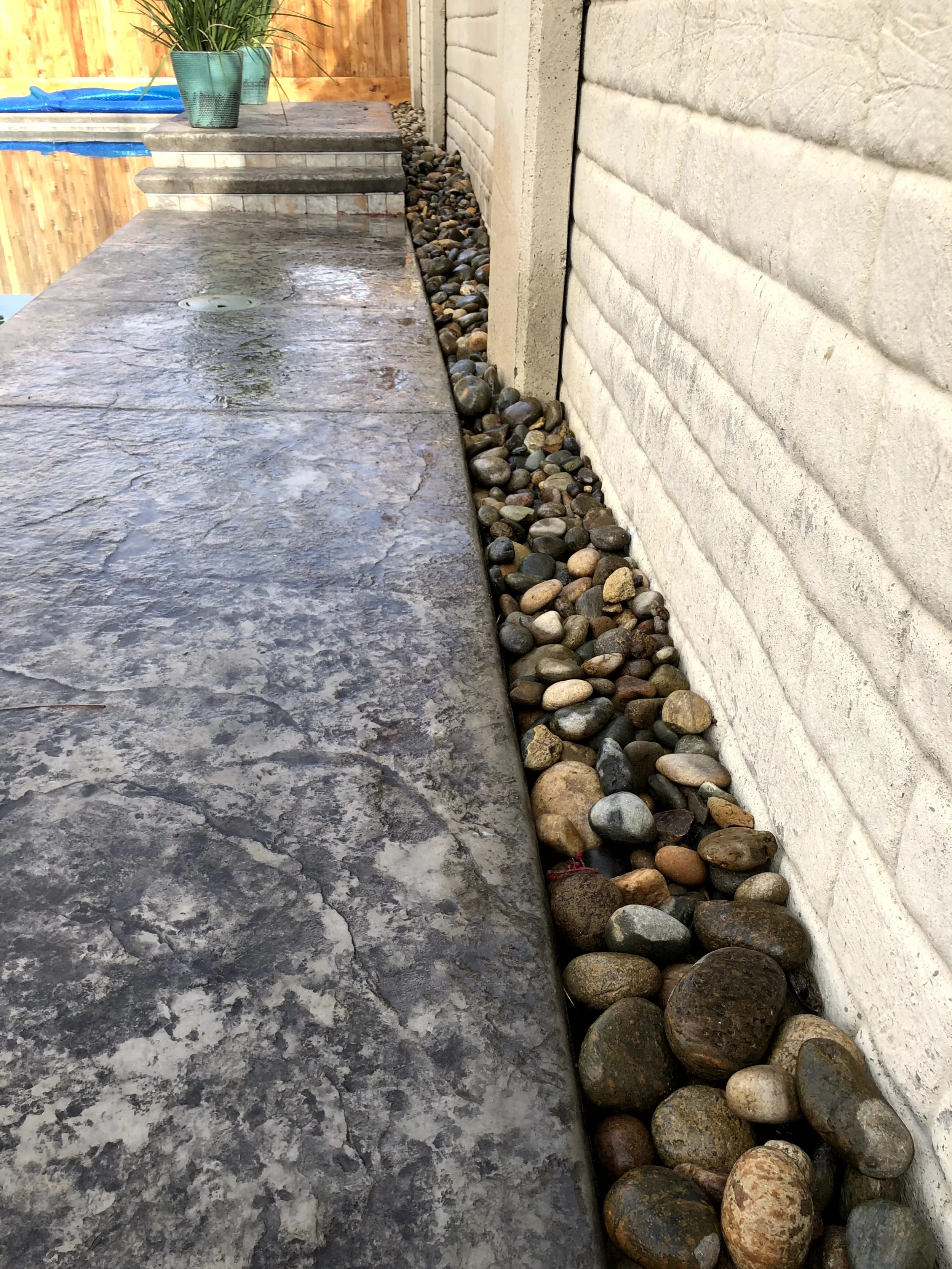 A stone-tiled patio with a small drainage hole, adjacent to a wall with rocks filling the gap at the base.