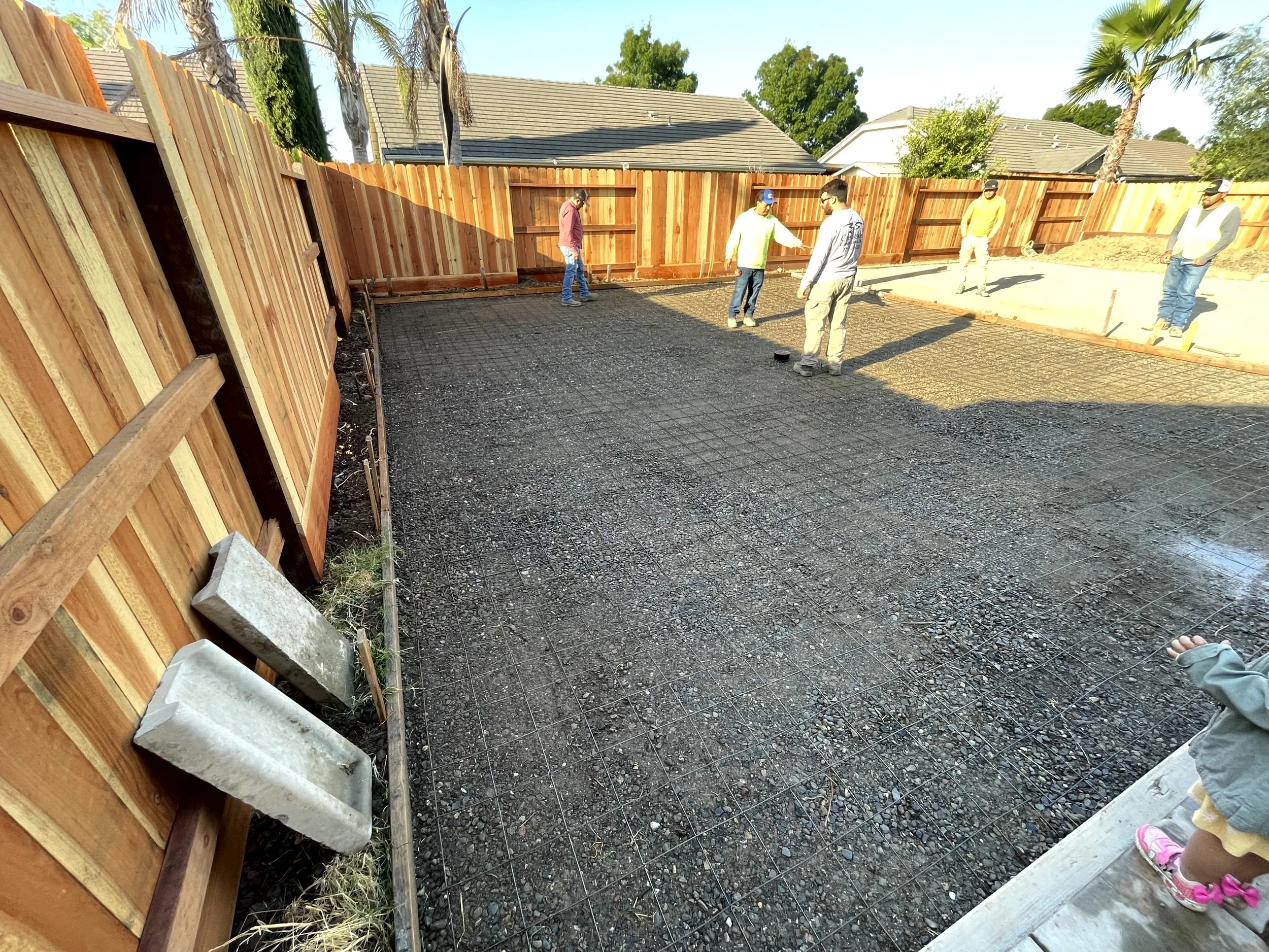 Construction workers pouring concrete on a backyard patio, with wooden fence and houses in the background.