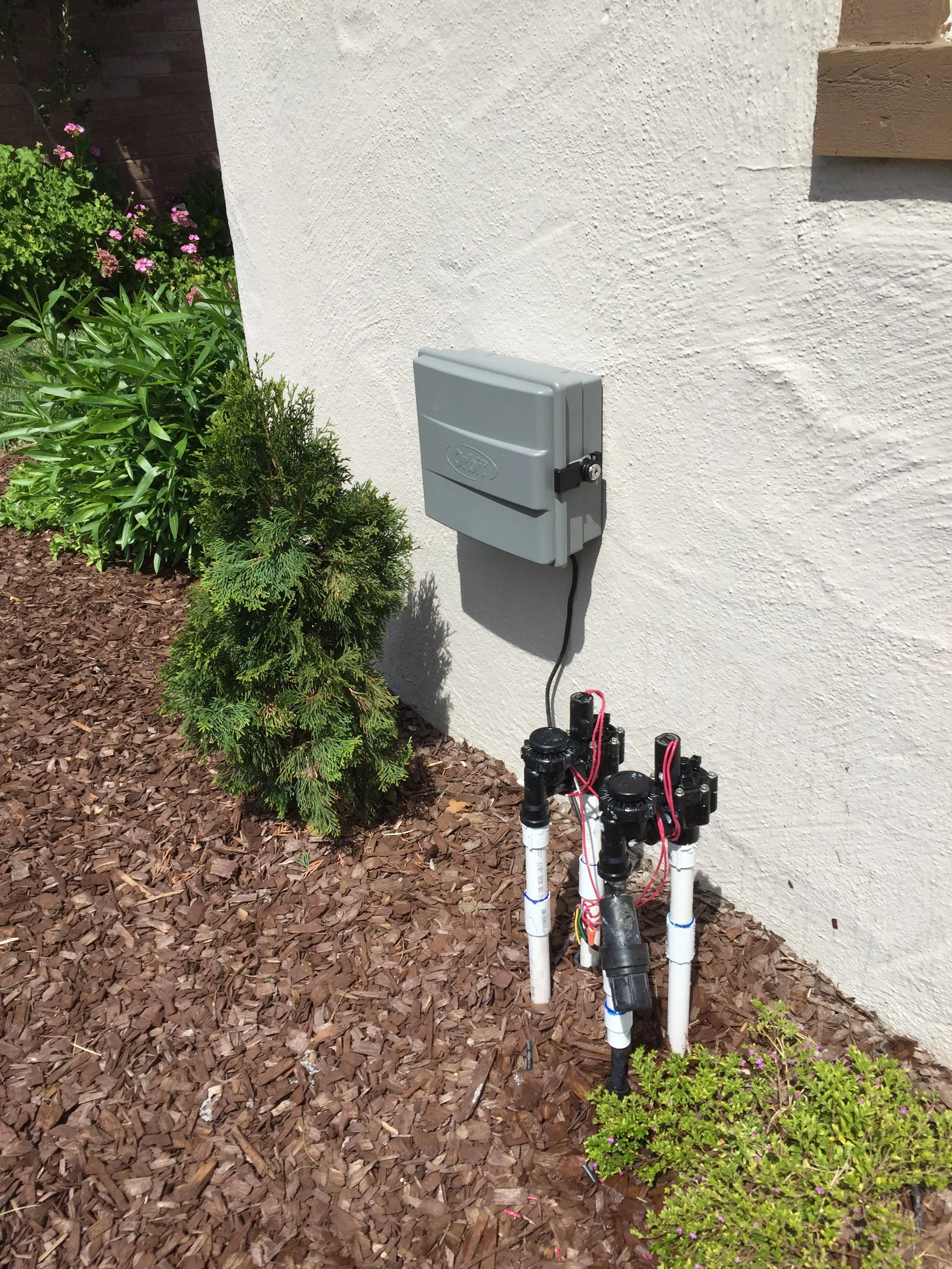 An outdoor scene showing an irrigation system with pipes and valves next to a white house wall, a gray electrical box mounted on the wall, and surrounding green plants and mulch.