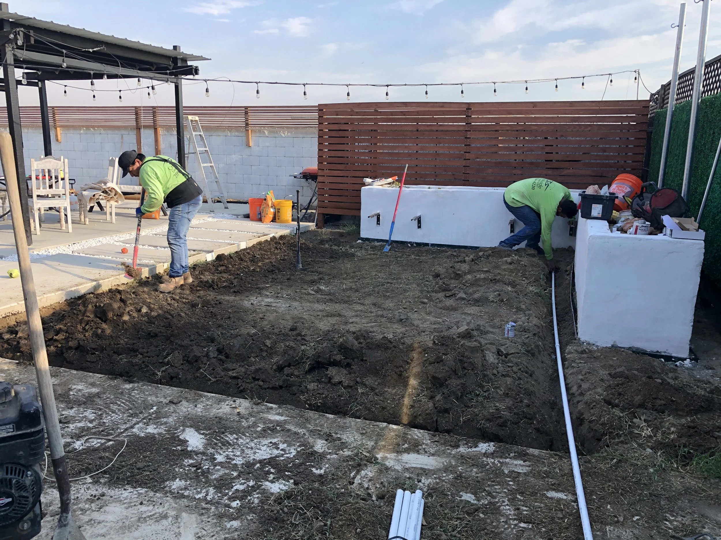 Two workers are laying down concrete or paving stones in a backyard patio area under a pergola, with construction tools and materials around them. One worker is using a tamper, and the other is working near a white structure.
