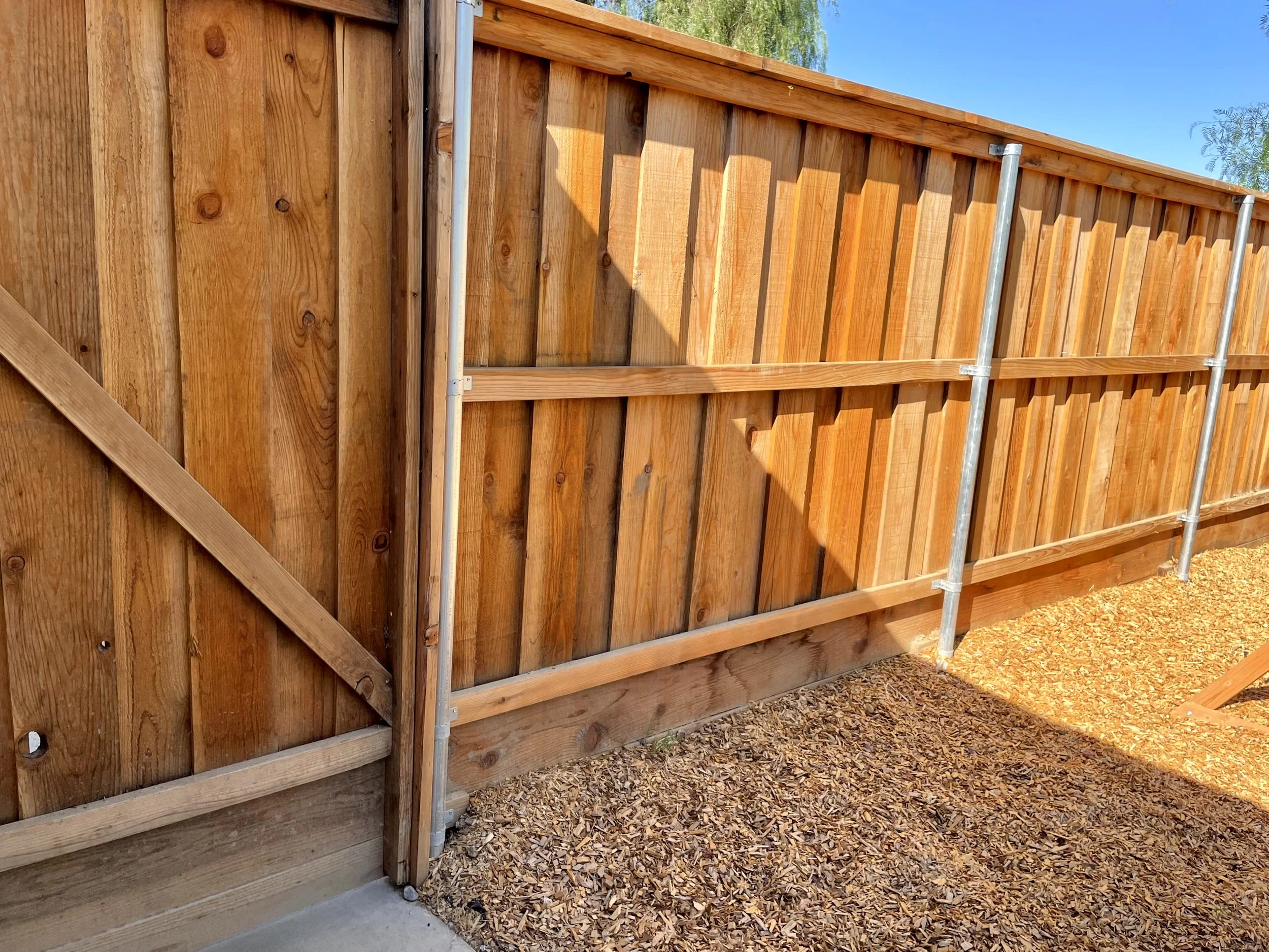A newly built wooden privacy fence with metal posts in a backyard, with the ground covered in wood mulch and a clear blue sky overhead.