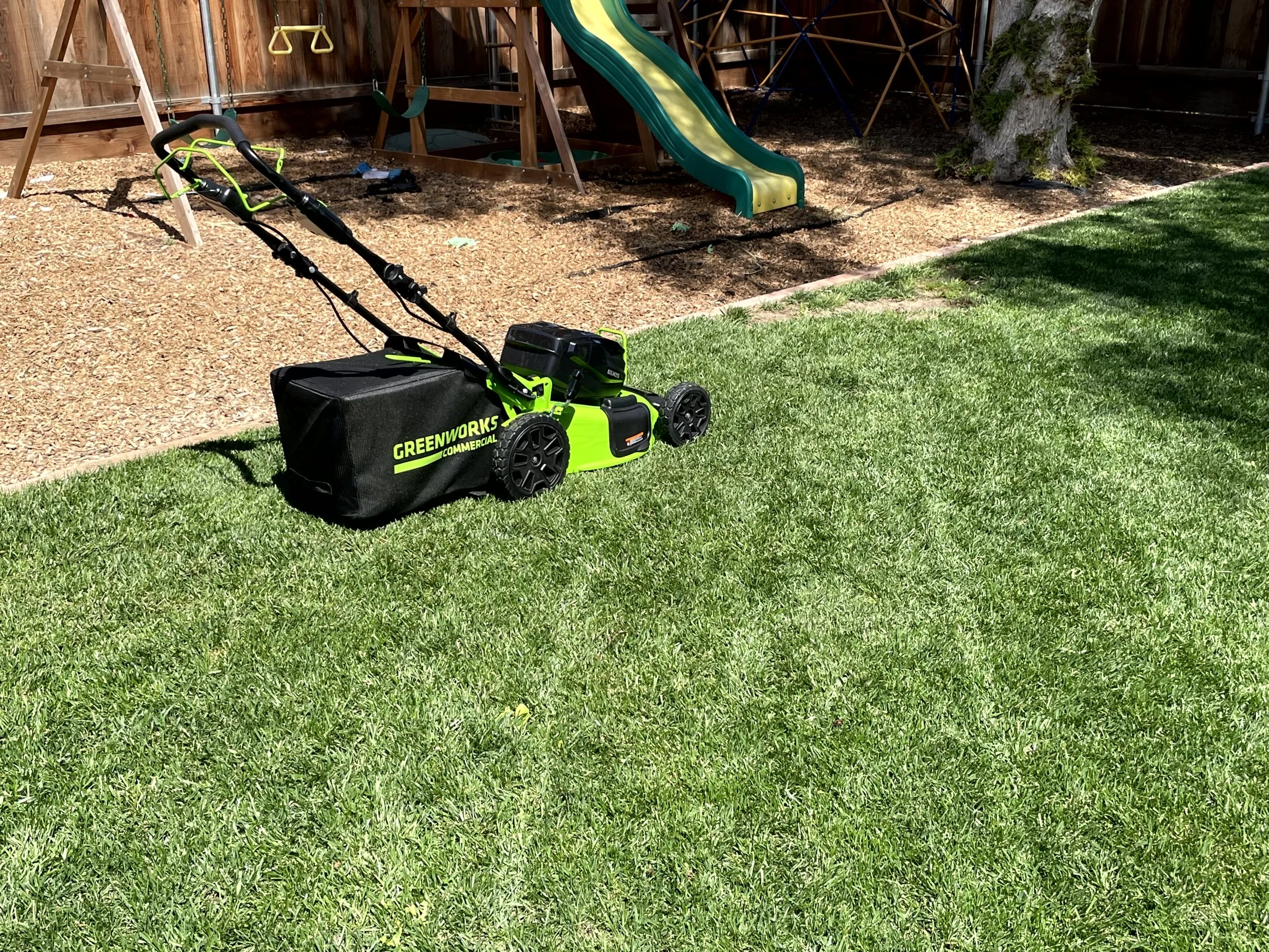 GreenWorks electric lawn mower on a well-manicured lawn, with a mulch bed and playground equipment including a slide and swings in the background.