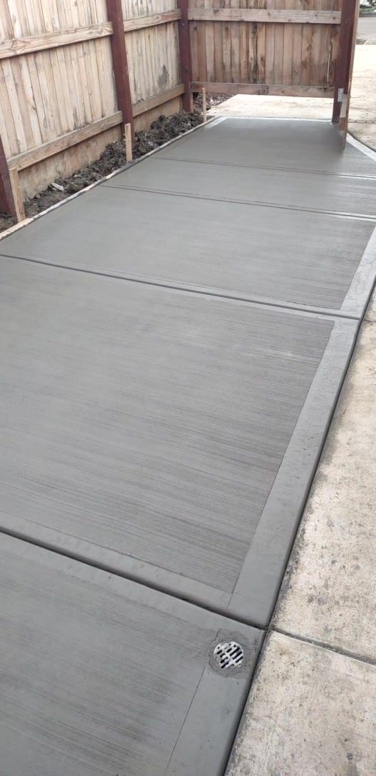 Newly poured concrete sidewalk with a circular drain in a residential area, bordered by a wooden fence.