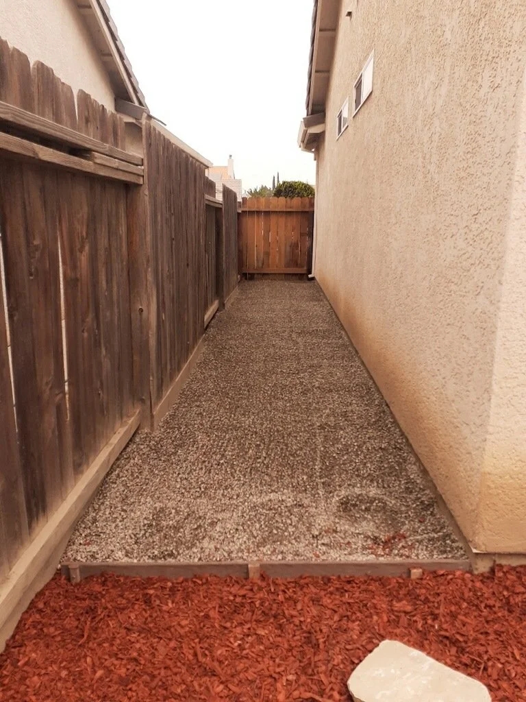 A narrow backyard pathway enclosed by a wooden fence on the left and a beige wall on the right, with recent gravel installation and red mulch at the foreground.