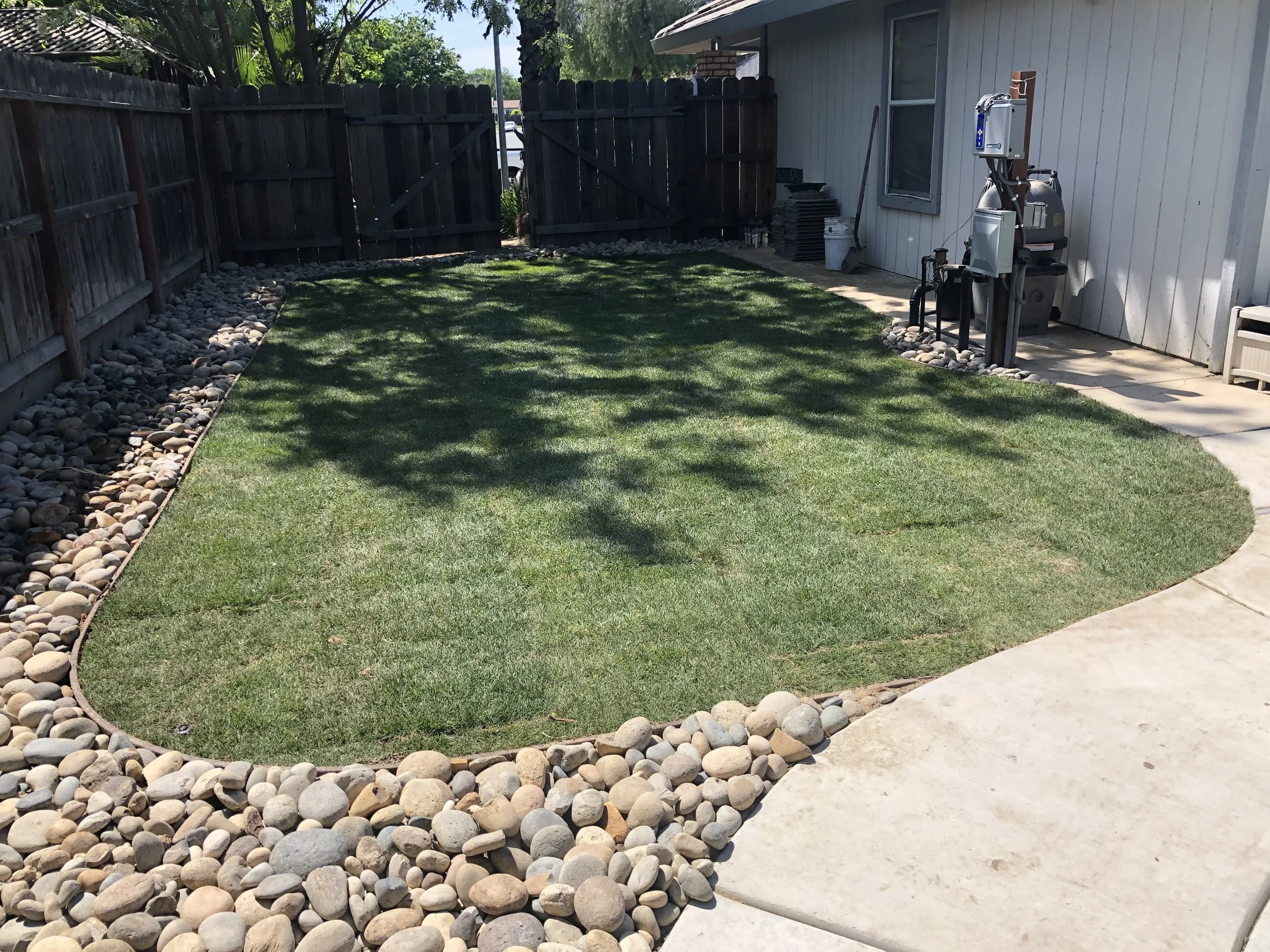 Backyard with a small grass lawn bordered by smooth river rocks, wooden fence, and a house with a concrete patio area, backyard grill, and tools. Shadows of trees cast on the grass.