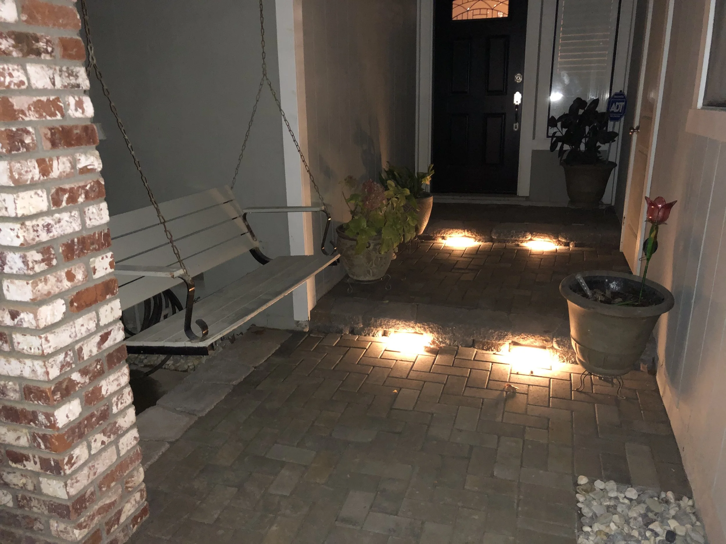 Nighttime view of a porch with potted plants, a hanging bench, brick and wooden flooring, and small lights illuminating the pathway.