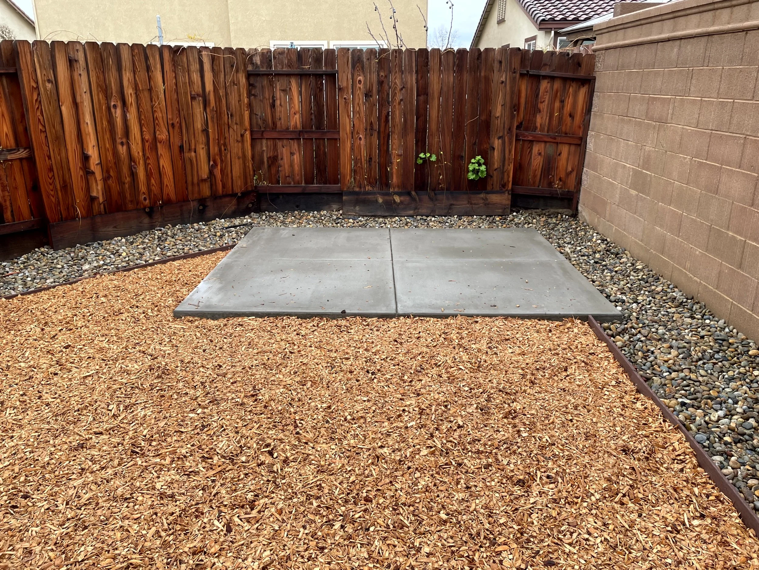 Backyard with a wooden fence, concrete patio, mulch bed, and gravel border on a cloudy day.