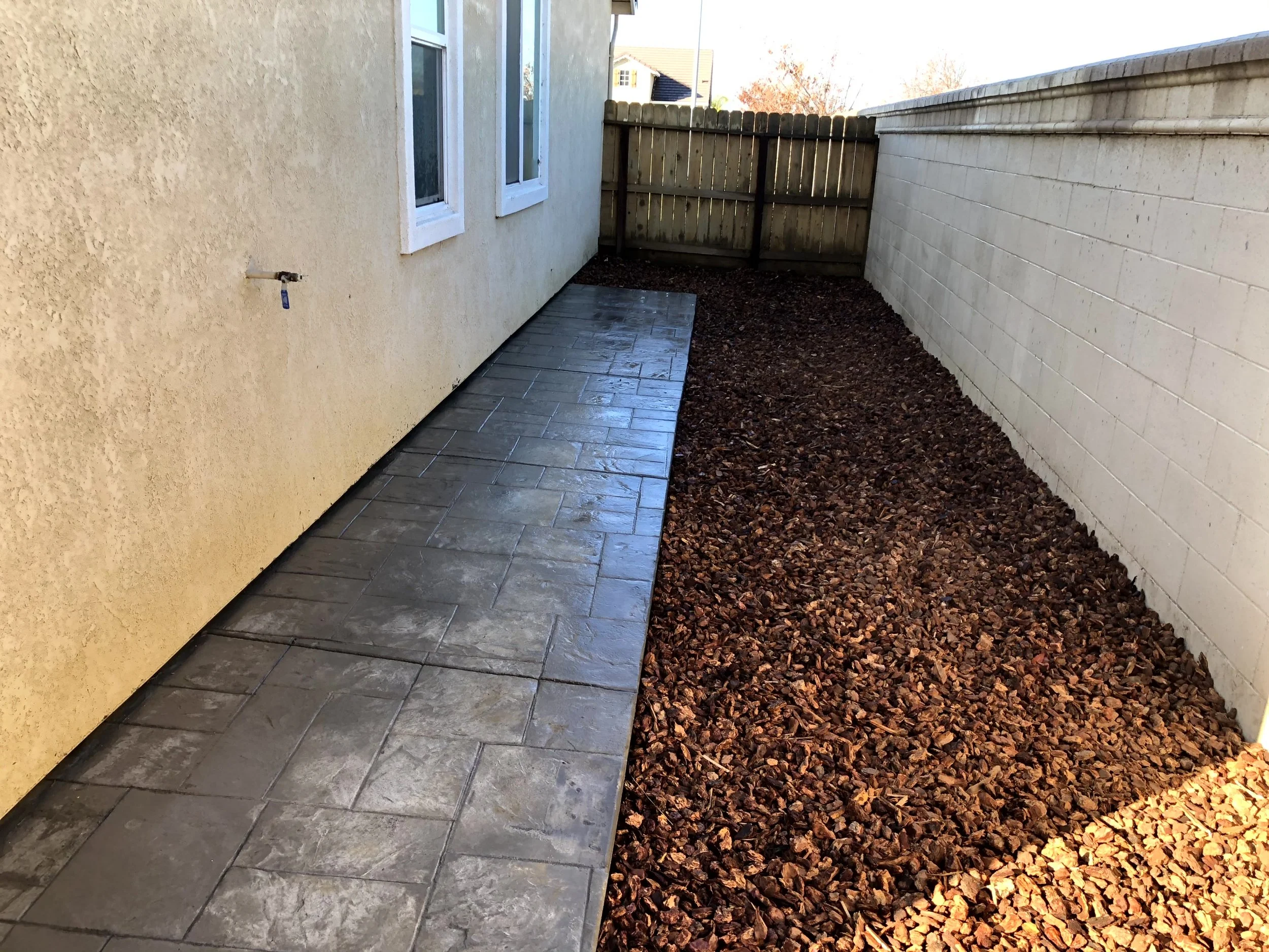 Photo of a narrow backyard with a newly paved concrete walkway on the left, a beige stucco house with three windows on the left, and a wooden fence at the back. The right side has a white brick wall, and there is reddish-brown mulch on the ground to