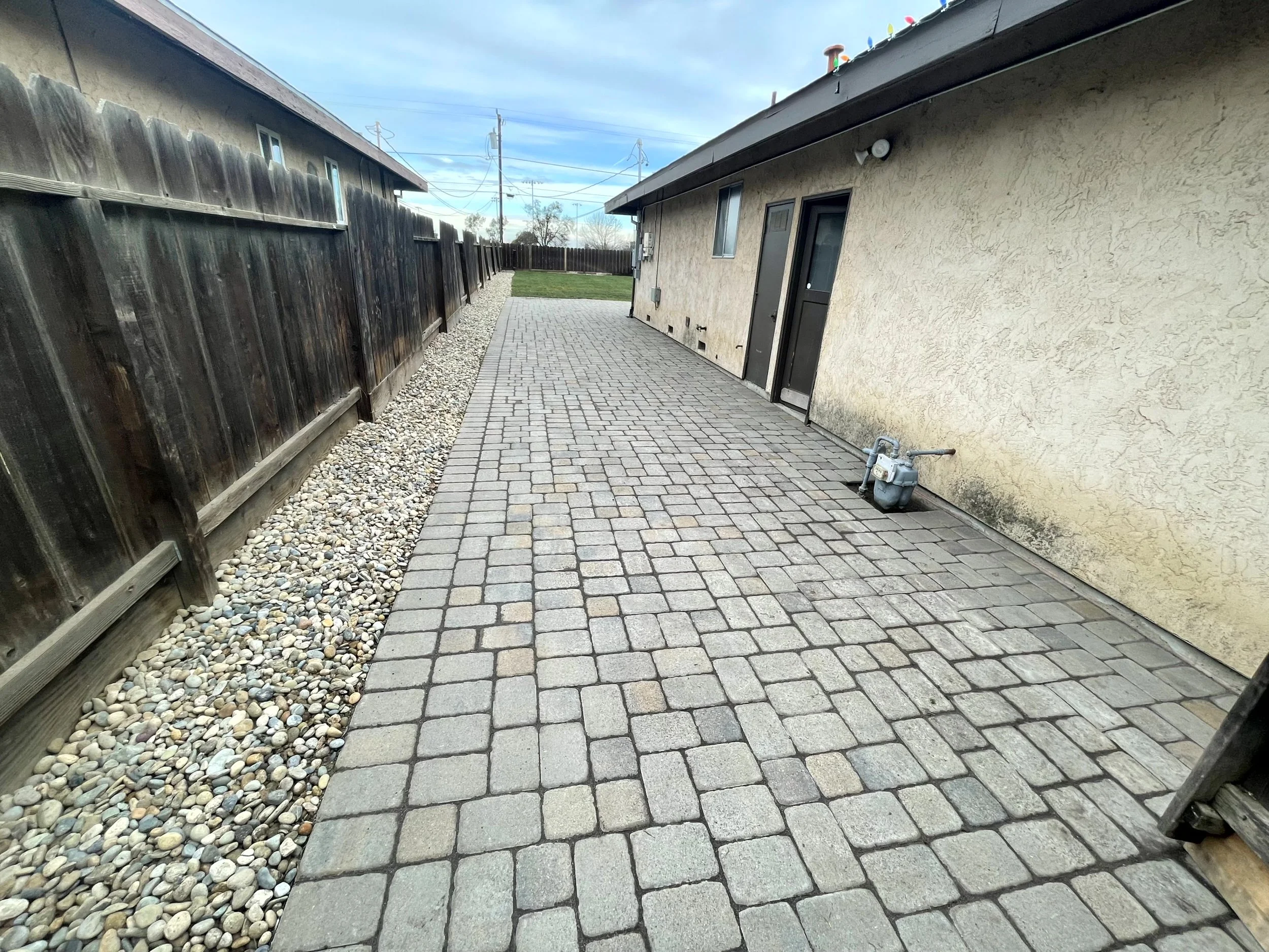 Backyard patio with brick pavers, a wooden fence on the left, and a beige house wall on the right, with a small grassy area in the background, under a partly cloudy sky.