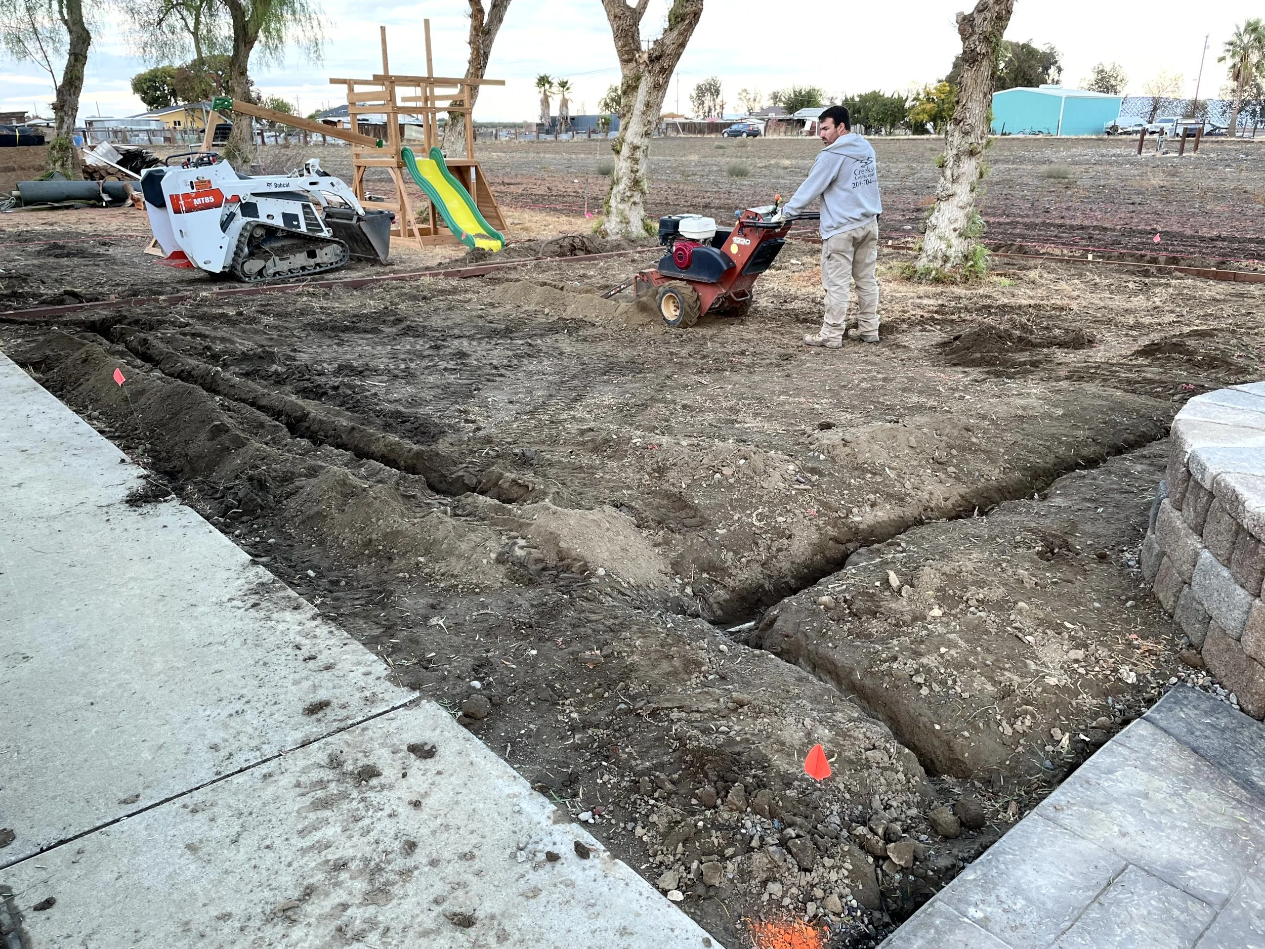 A construction site shows a person operating a mini compactor on dirt, with an outdoor playset and a skid steer loader nearby, and trees in the background.