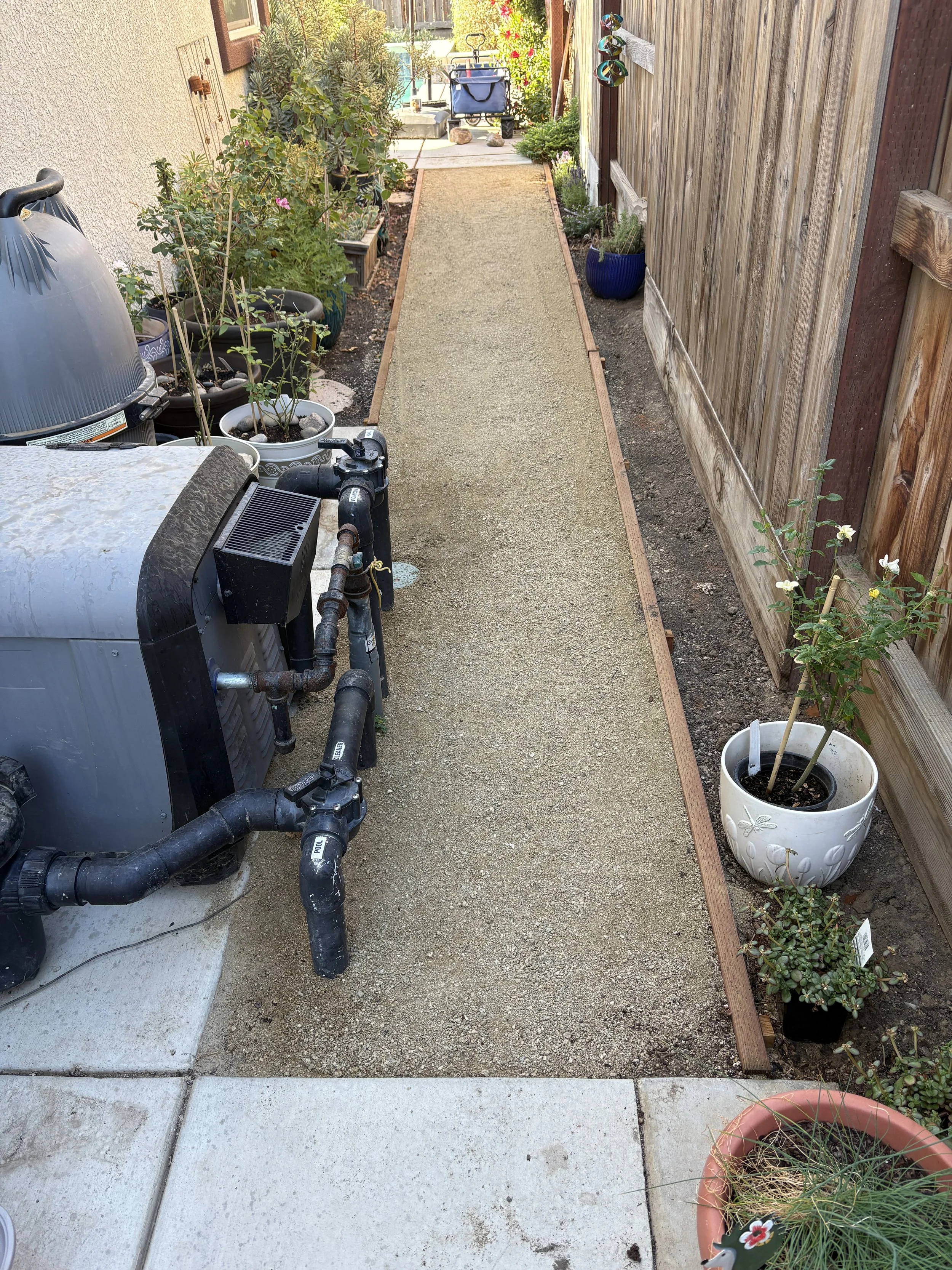 A small backyard pathway with a gravel strip bordered by wood edging, set between a concrete patio and a wooden fence, with potted plants and gardening equipment along the sides.