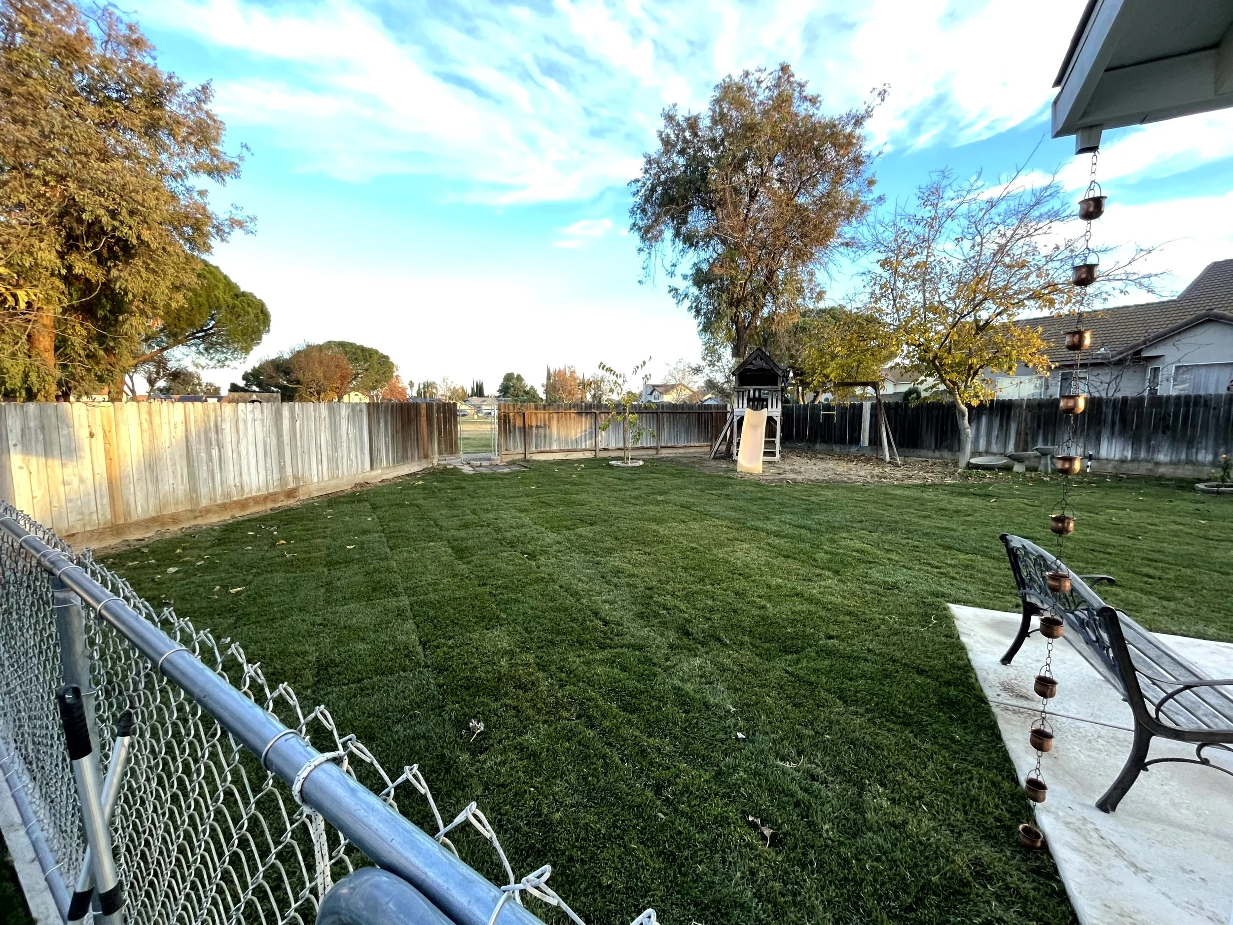 A backyard with a grassy lawn, a wooden fence, a swing set with a slide, a tree with autumn leaves, a bench, and a wind chime hanging from the house eaves, with a cloudy sky overhead.