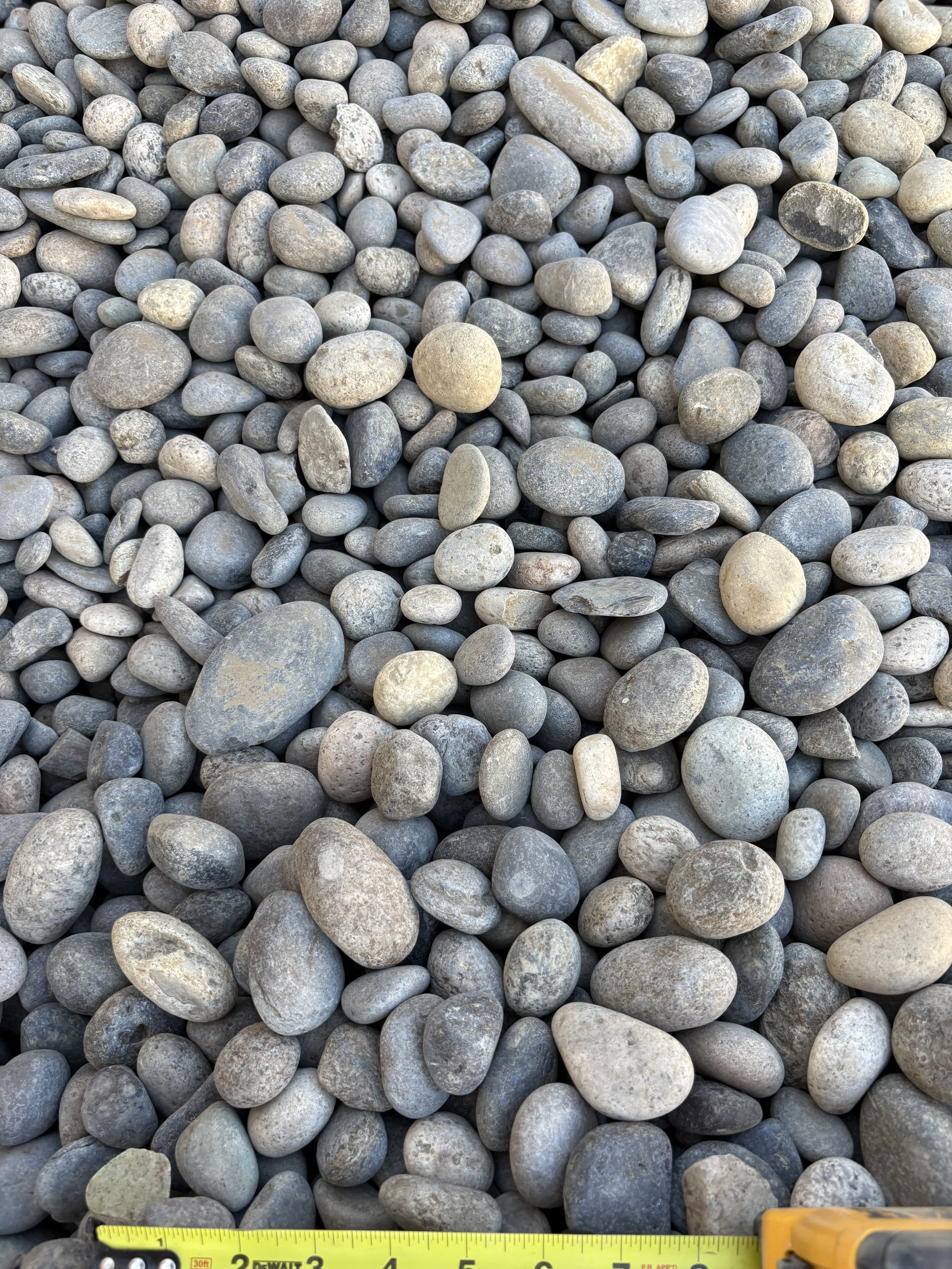 A collection of various-sized smooth, rounded gray and beige river rocks, with a yellow measuring tape at the bottom showing inches.