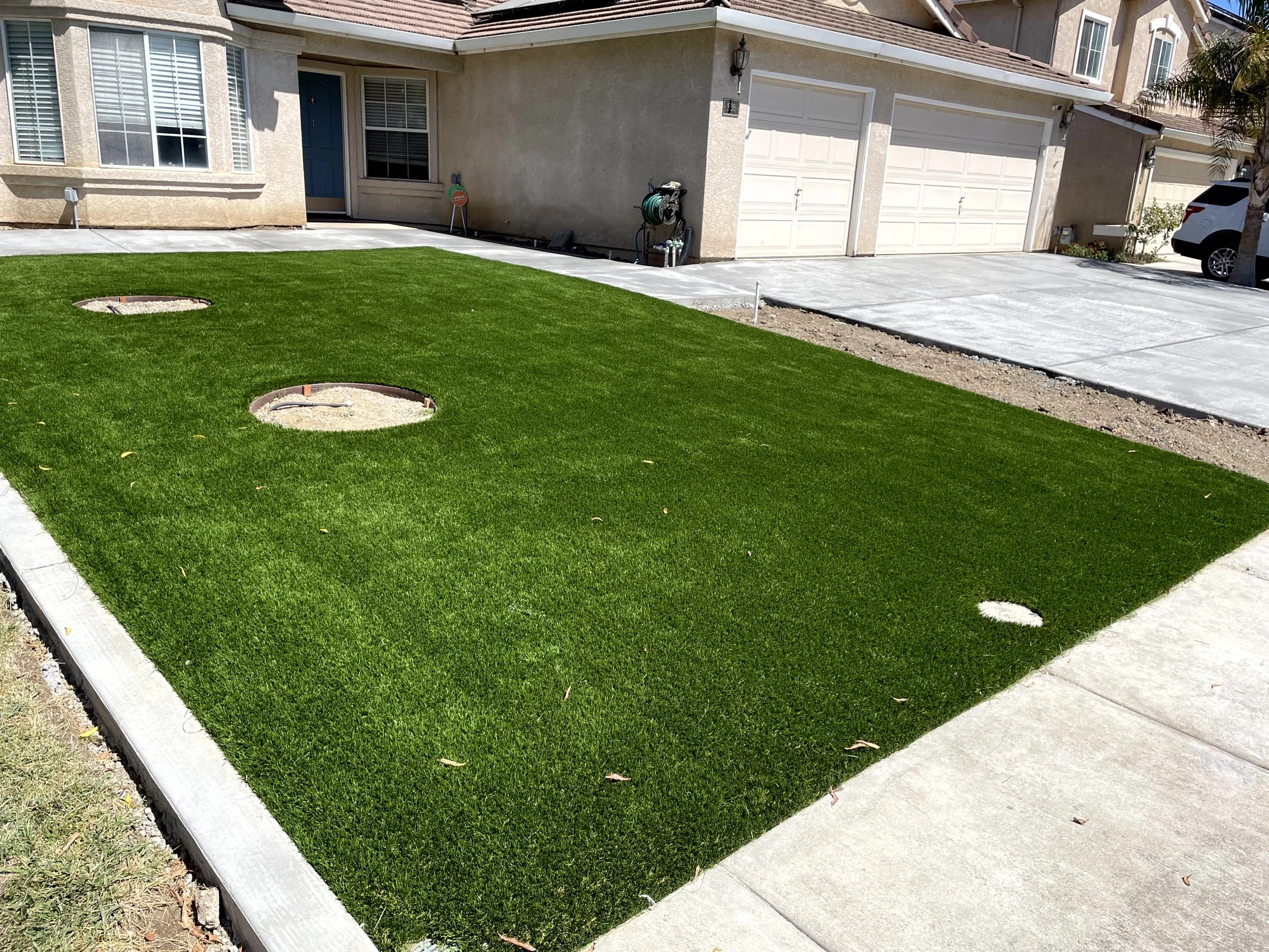 A newly landscaped front yard with bright green grass, in-ground sprinkler system, and a concrete driveway leading to a two-car garage in a suburban neighborhood.