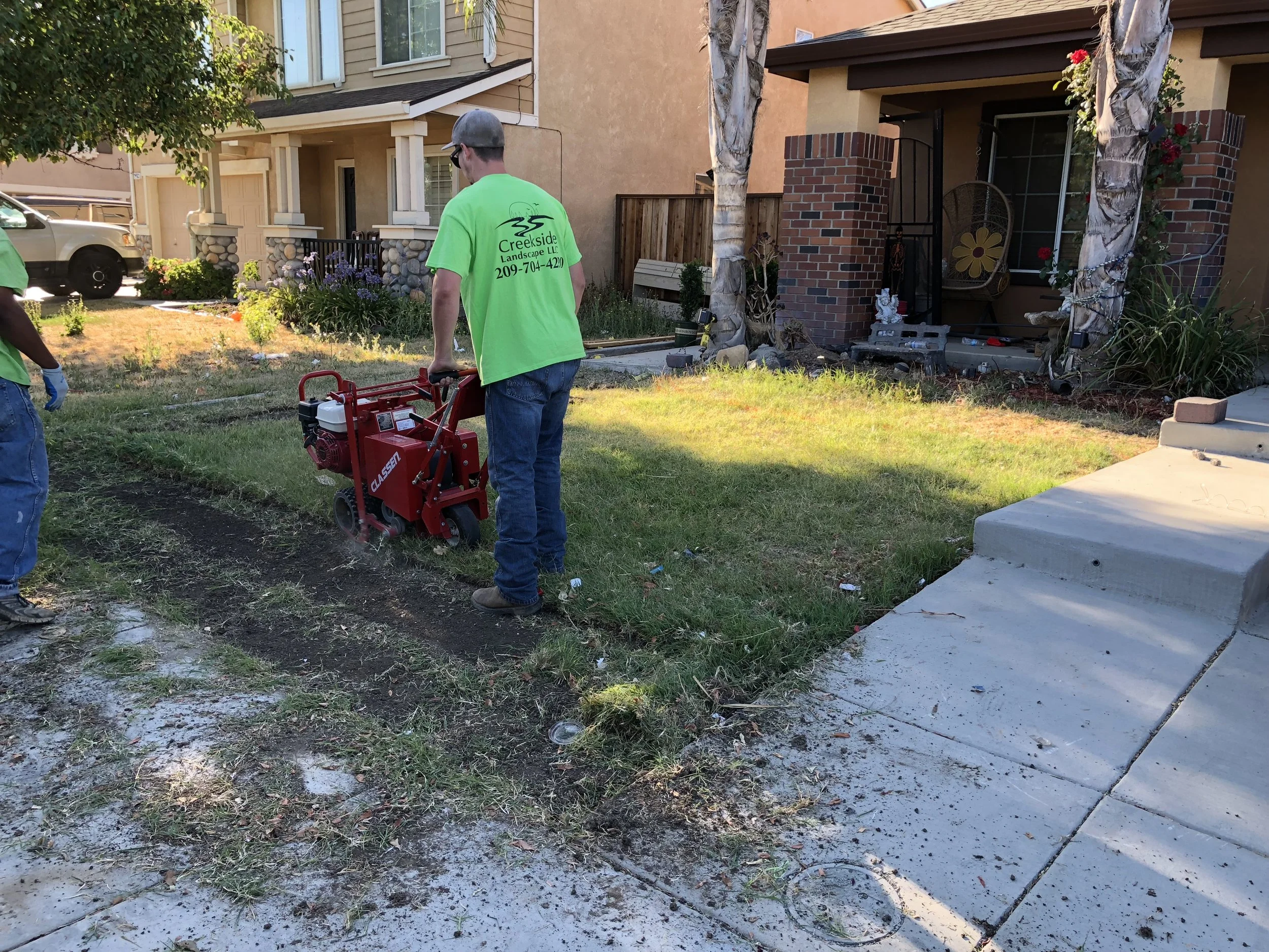 A worker in a bright green shirt operating a sod cutter on a lawn in front of a house.