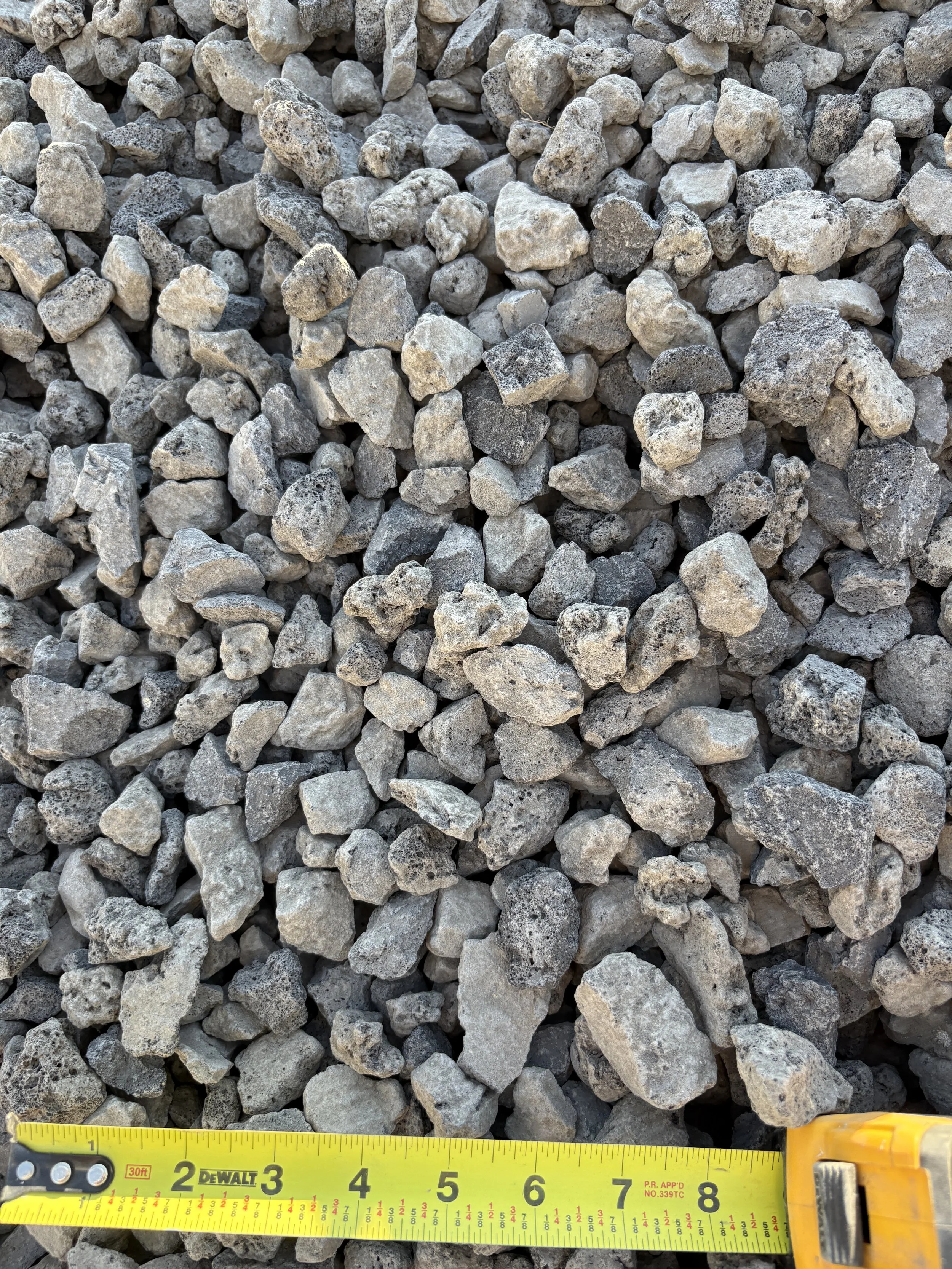 A close-up view of small, irregularly shaped gray gravel stones with a yellow measuring tape showing approximately 8 inches at the bottom of the image.