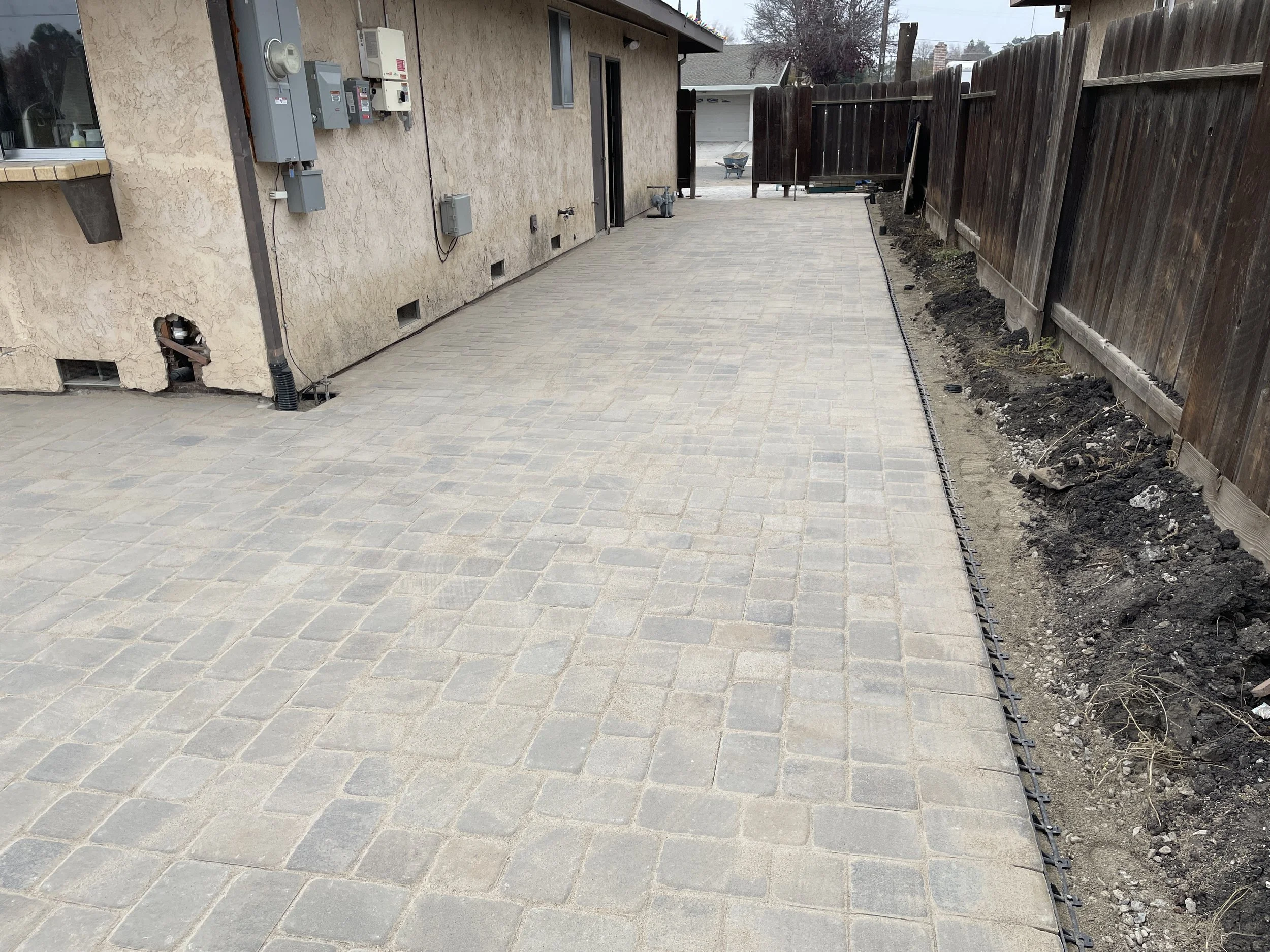 Newly paved backyard with interlocking stone patio, unfinished garden bed along the fence, and utility meters on the house wall.