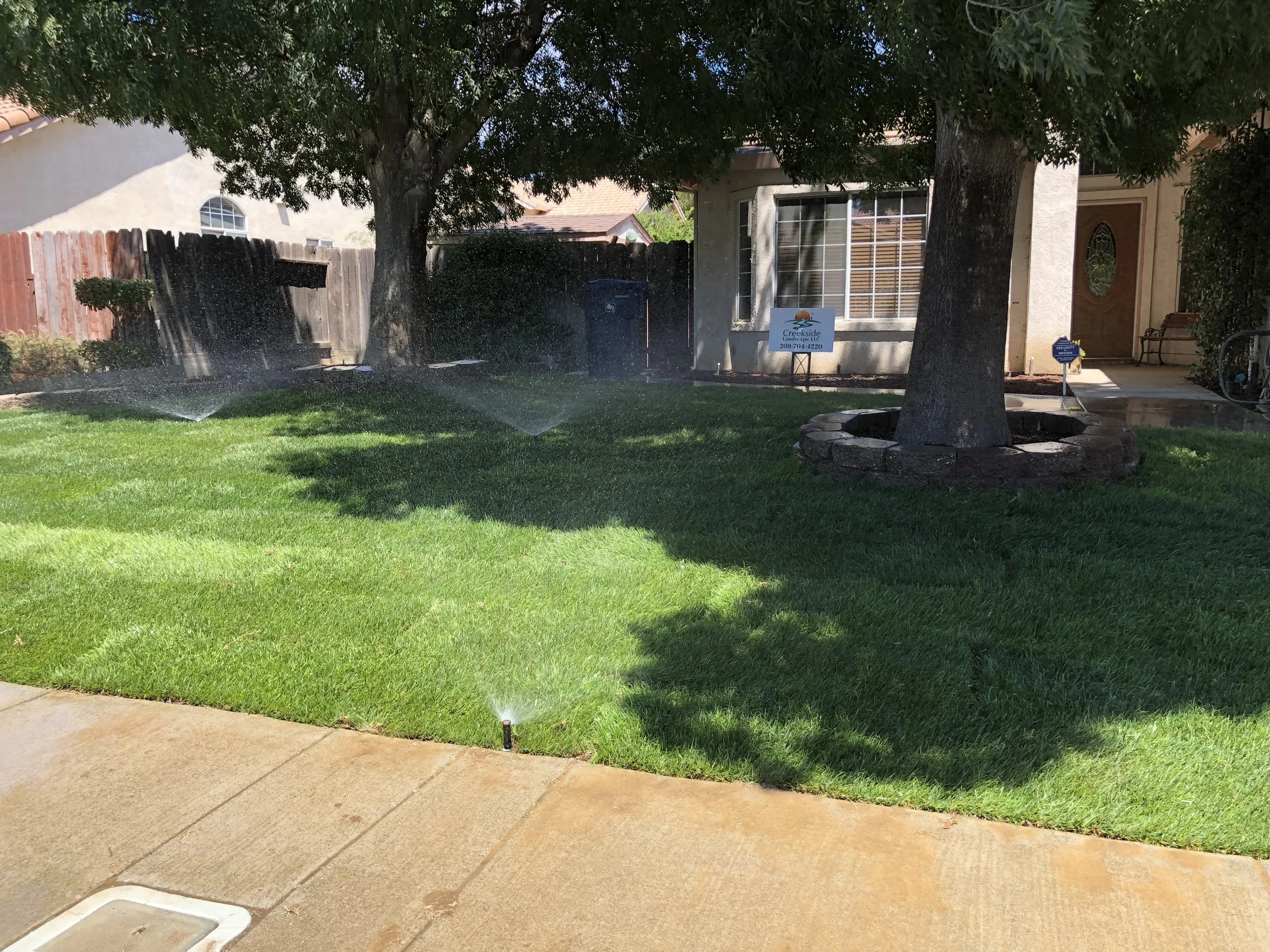 A well-maintained front yard with lush green grass being watered by sprinklers, shaded by large trees, and a house with a window, door, and a sign indicating a landscaping service.