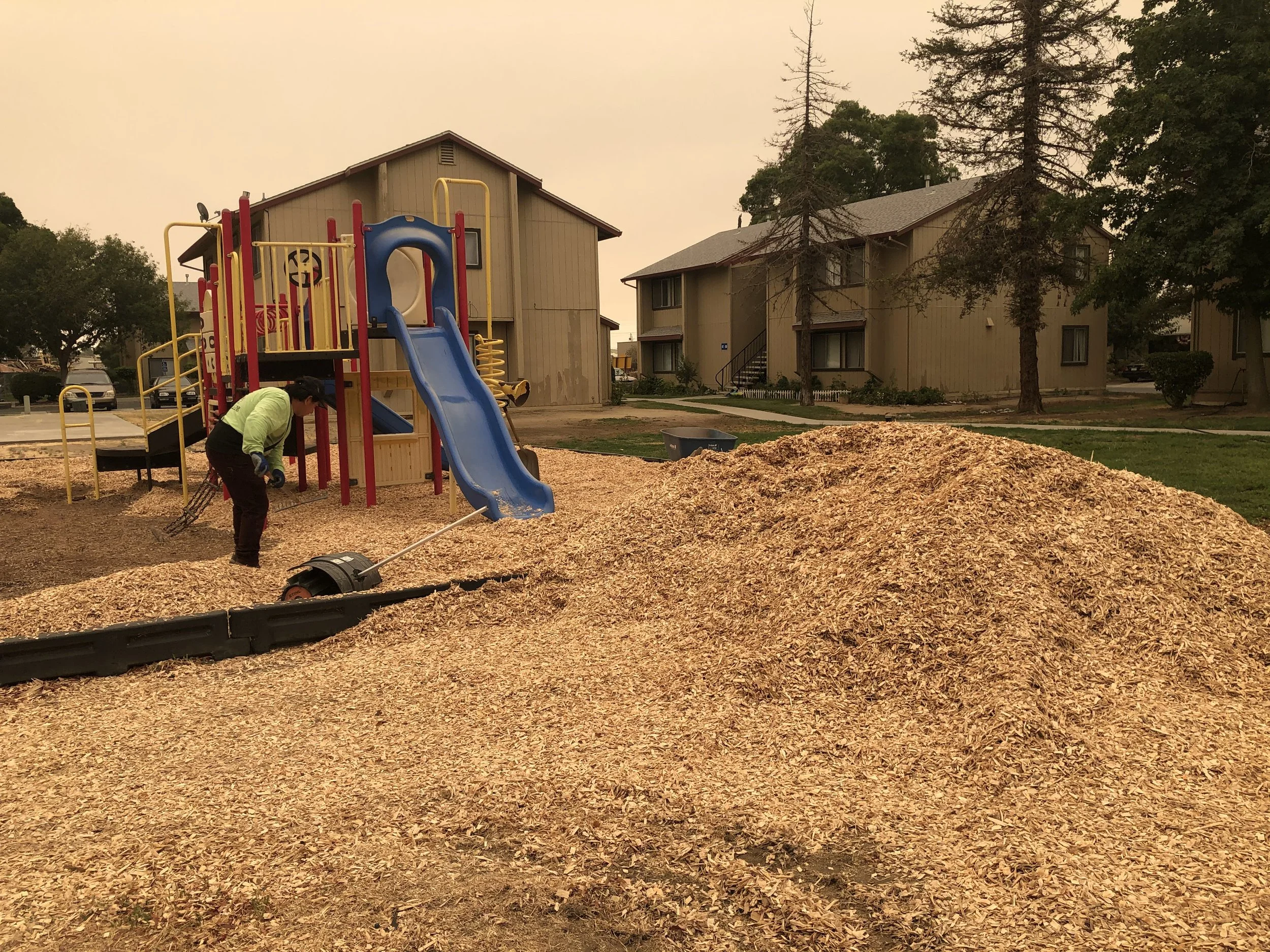 A person spreading mulch in a playground area with a blue slide, surrounded by houses and trees, during smoky weather.