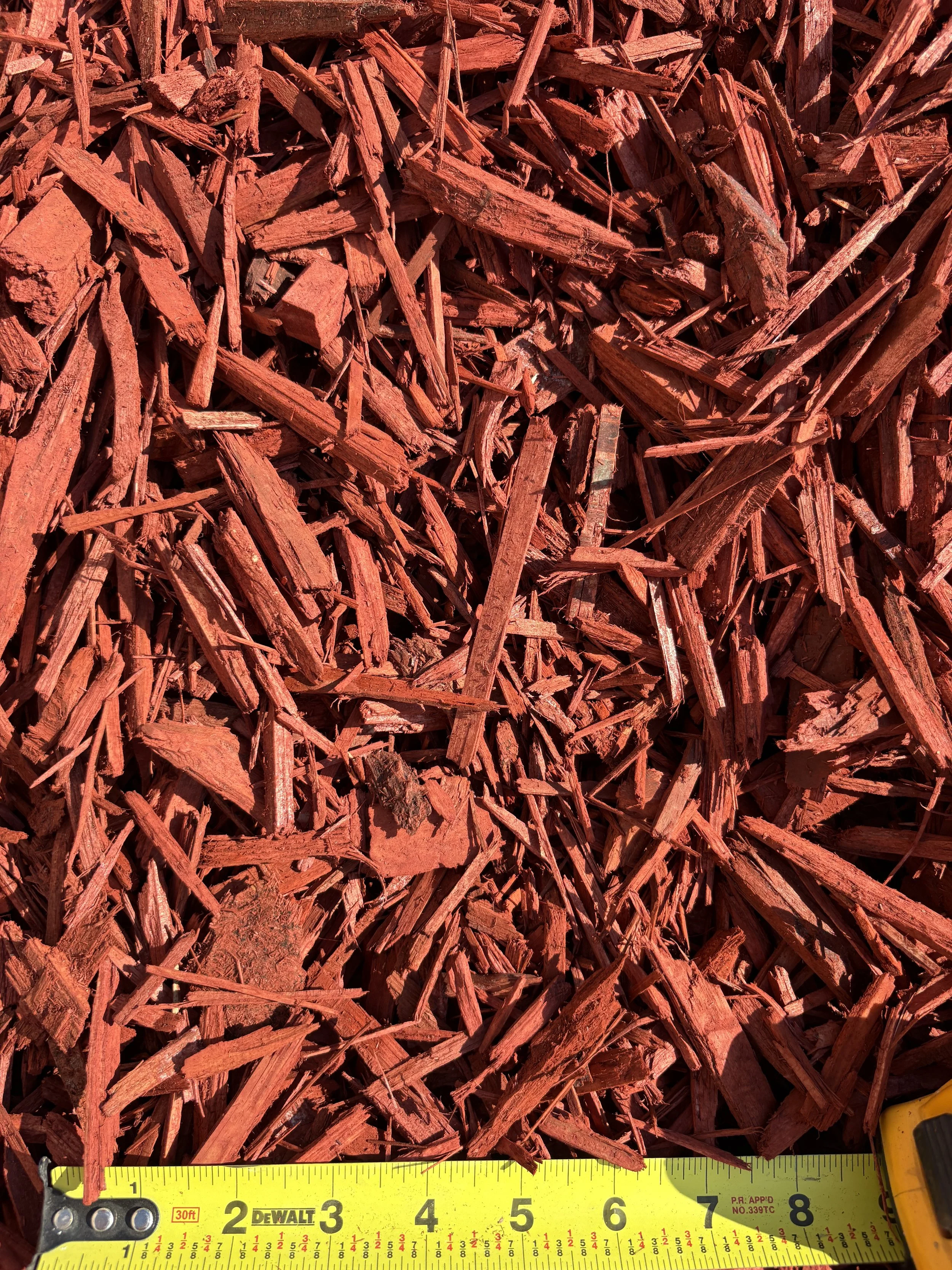 Close-up of red wood mulch with a yellow measuring tape at the bottom showing measurements from 1 to 8 inches.
