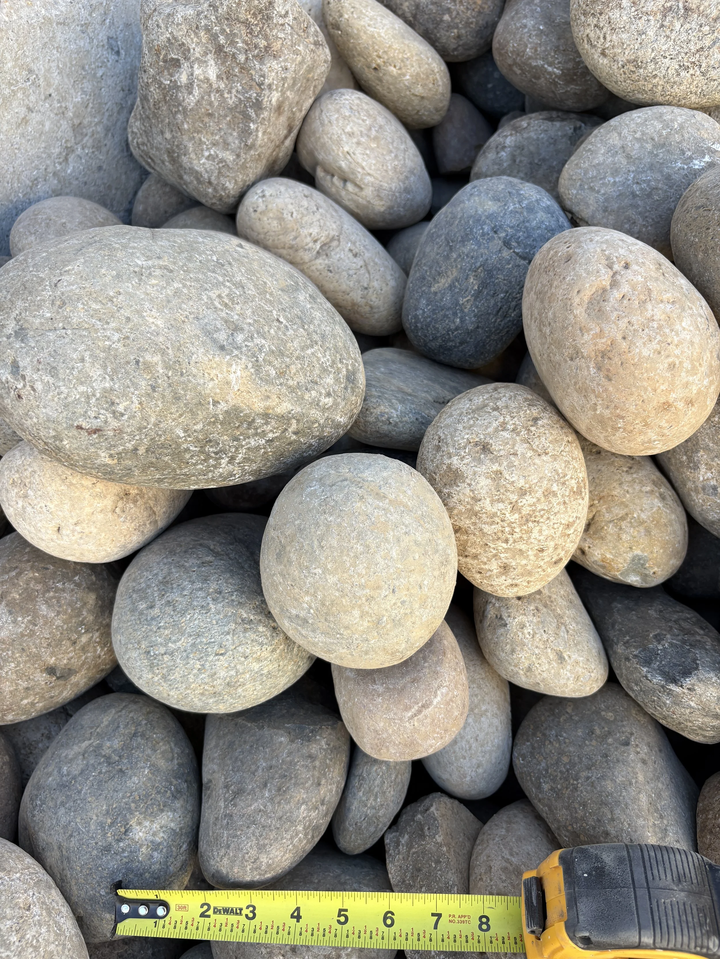 Close-up of various smooth, rounded river or beach stones of different sizes and colors, with a yellow measuring tape at the bottom for scale.