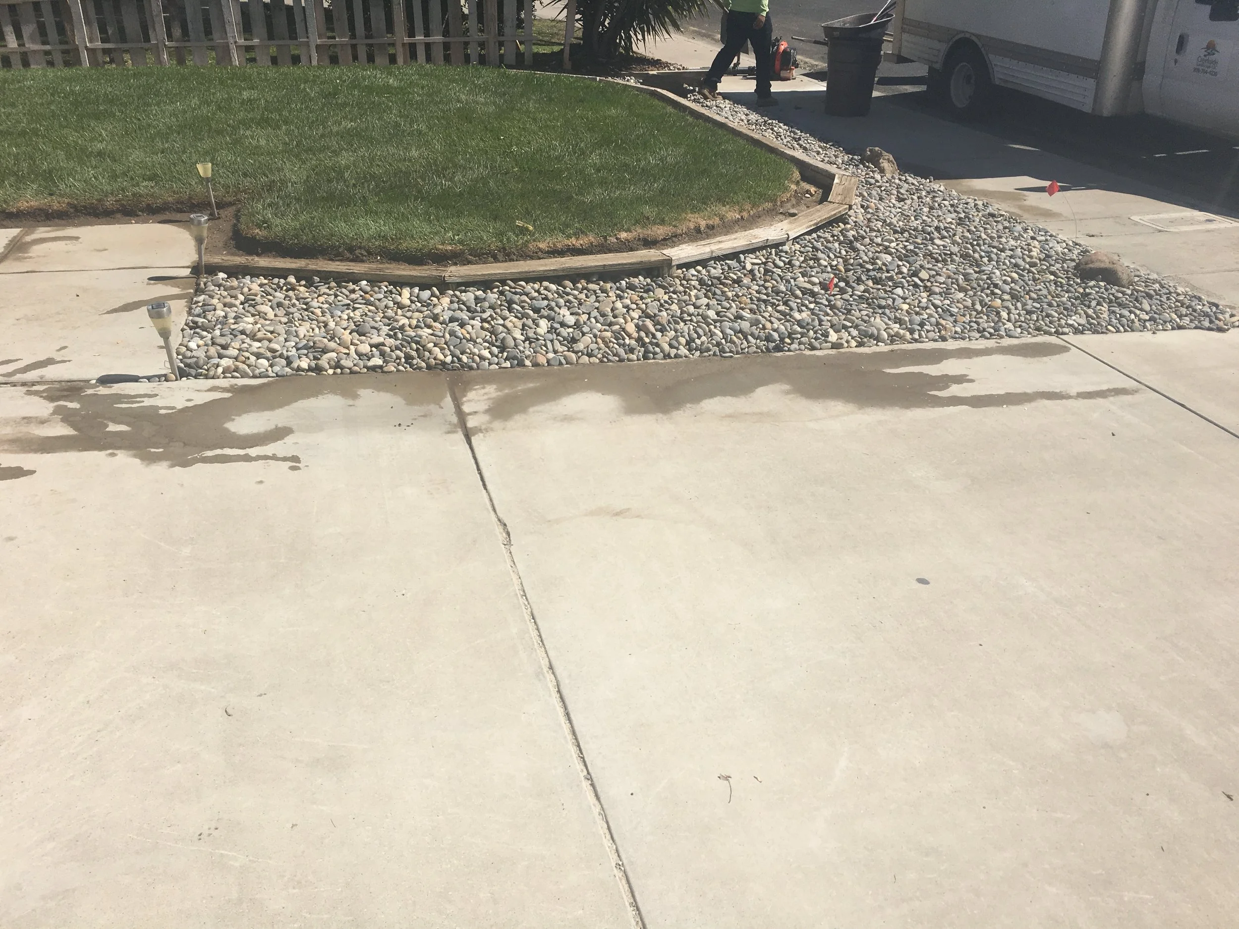 Front yard with small patch of grass, bordered by wood, and filled with rocks, adjacent to a paved sidewalk. There are landscape lights around the grass patch, with one light leaning. A person is visible in the background near a van, which has a tras