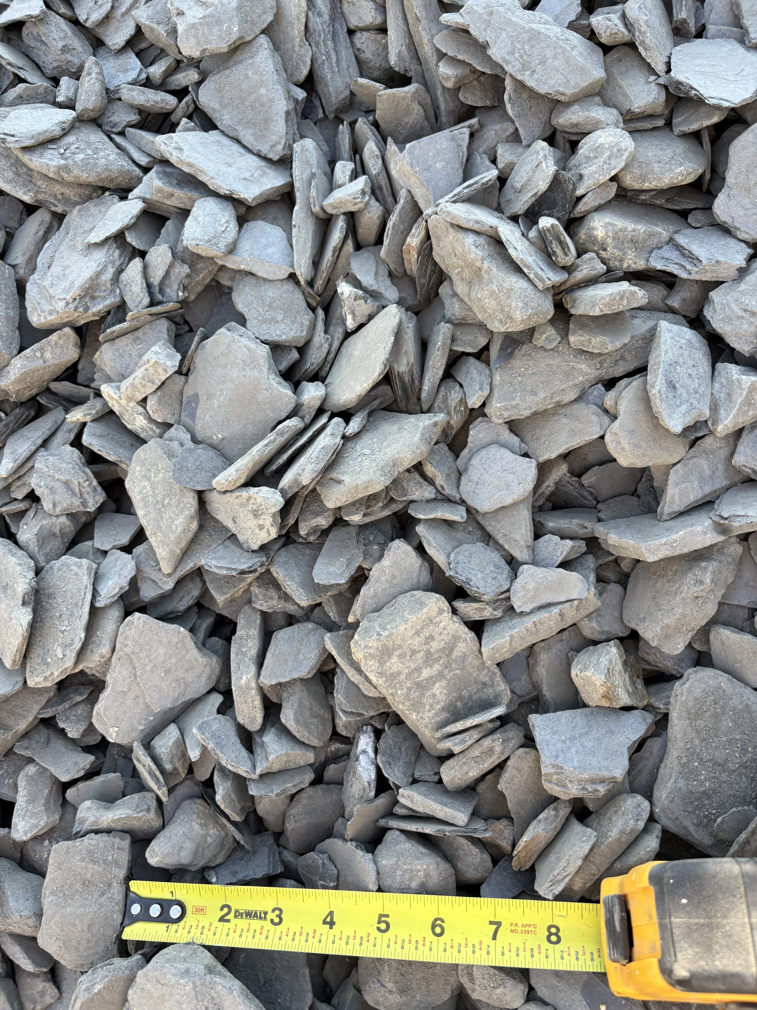 Close-up of various gray stones and rocks, with a yellow measuring tape showing approximately six inches in length at the bottom.