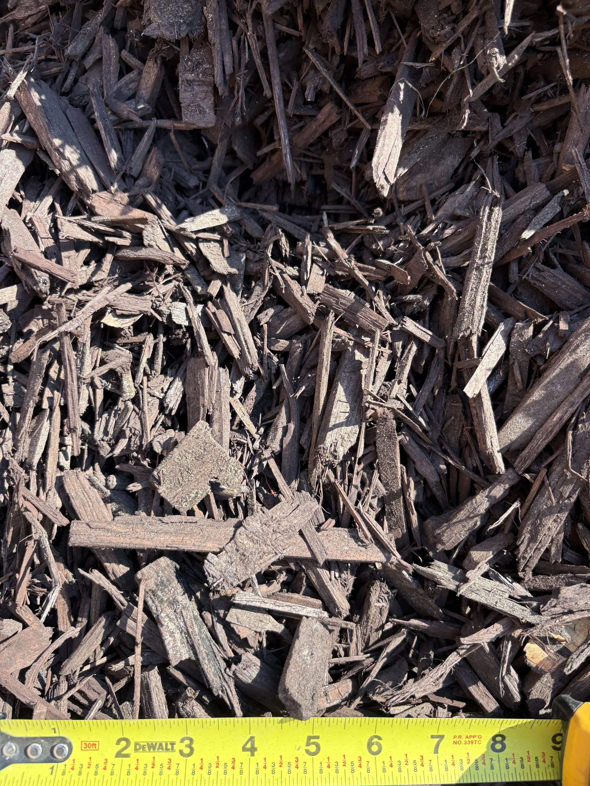 Close-up of dark brown shredded wood mulch with a yellow measuring tape at the bottom showing about 9 inches.