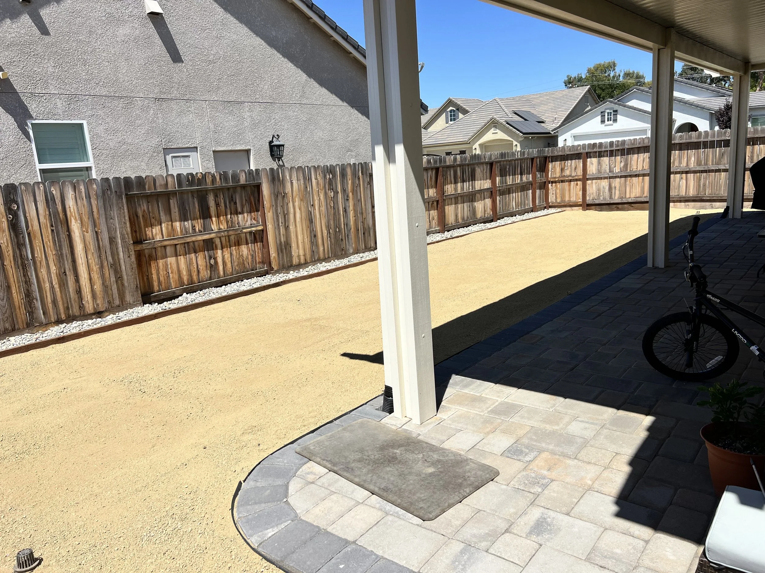 Backyard patio with a bike parked under a covered area, a small potted plant, and a wooden fence enclosing a yard with sand or gravel ground.