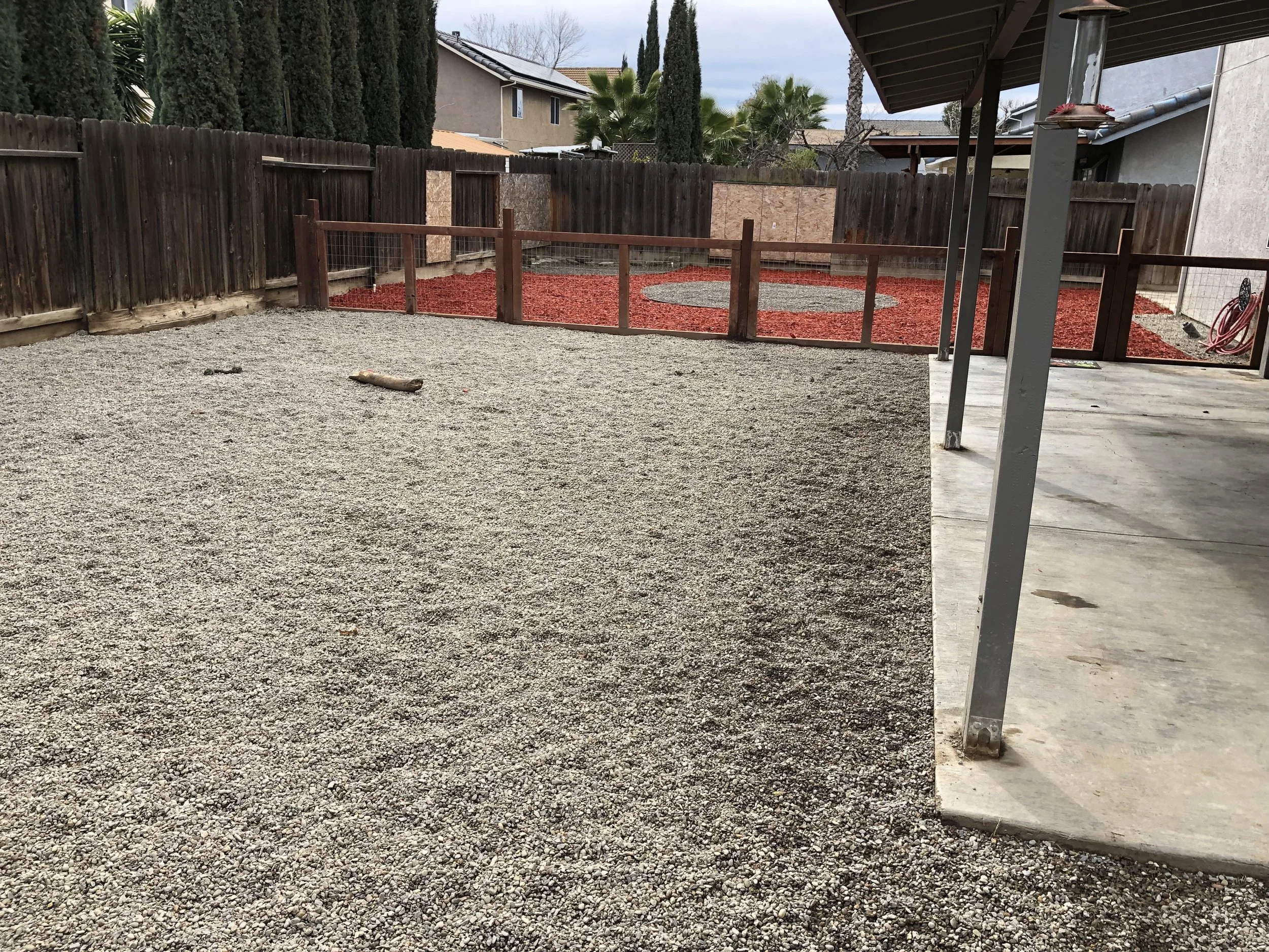 Backyard with a gravel area, a wooden fence with wire panels, and a red mulch area with a circular design. There is a covered porch with metal posts, and houses with trees are visible in the background.