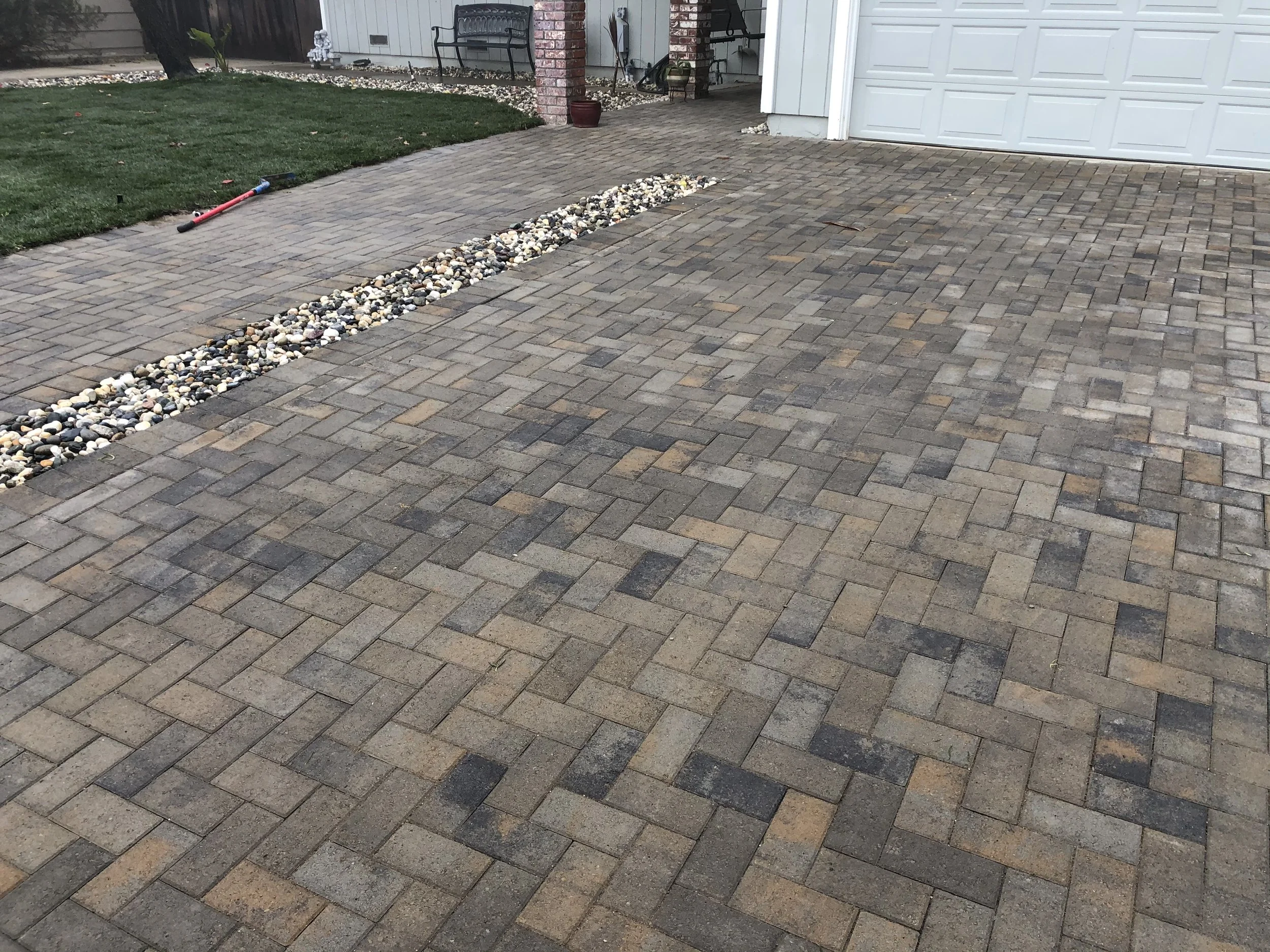 Newly paved brick driveway with a rock border separating it from a grassy yard. The driveway leads to a garage door, and the yard has a toy rake and a couple of chairs near the house.