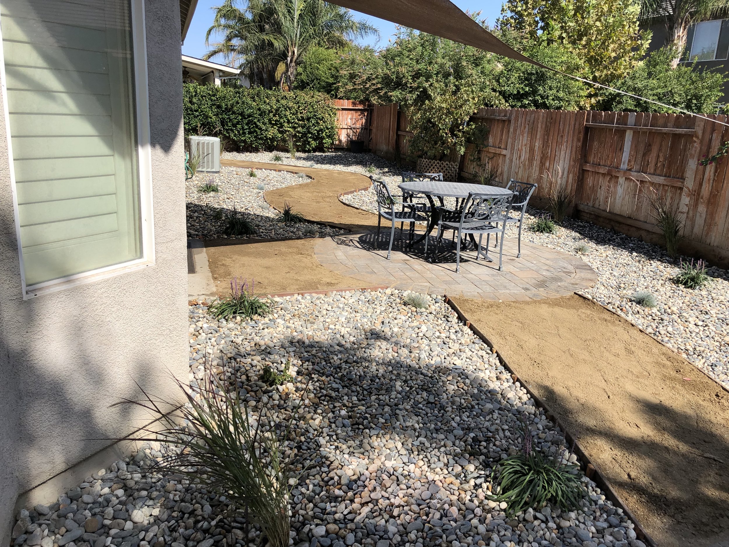 Backyard with a stone patio, outdoor table and chairs, landscaped with gravel and plants, enclosed by a wooden fence, with a curving pathway.