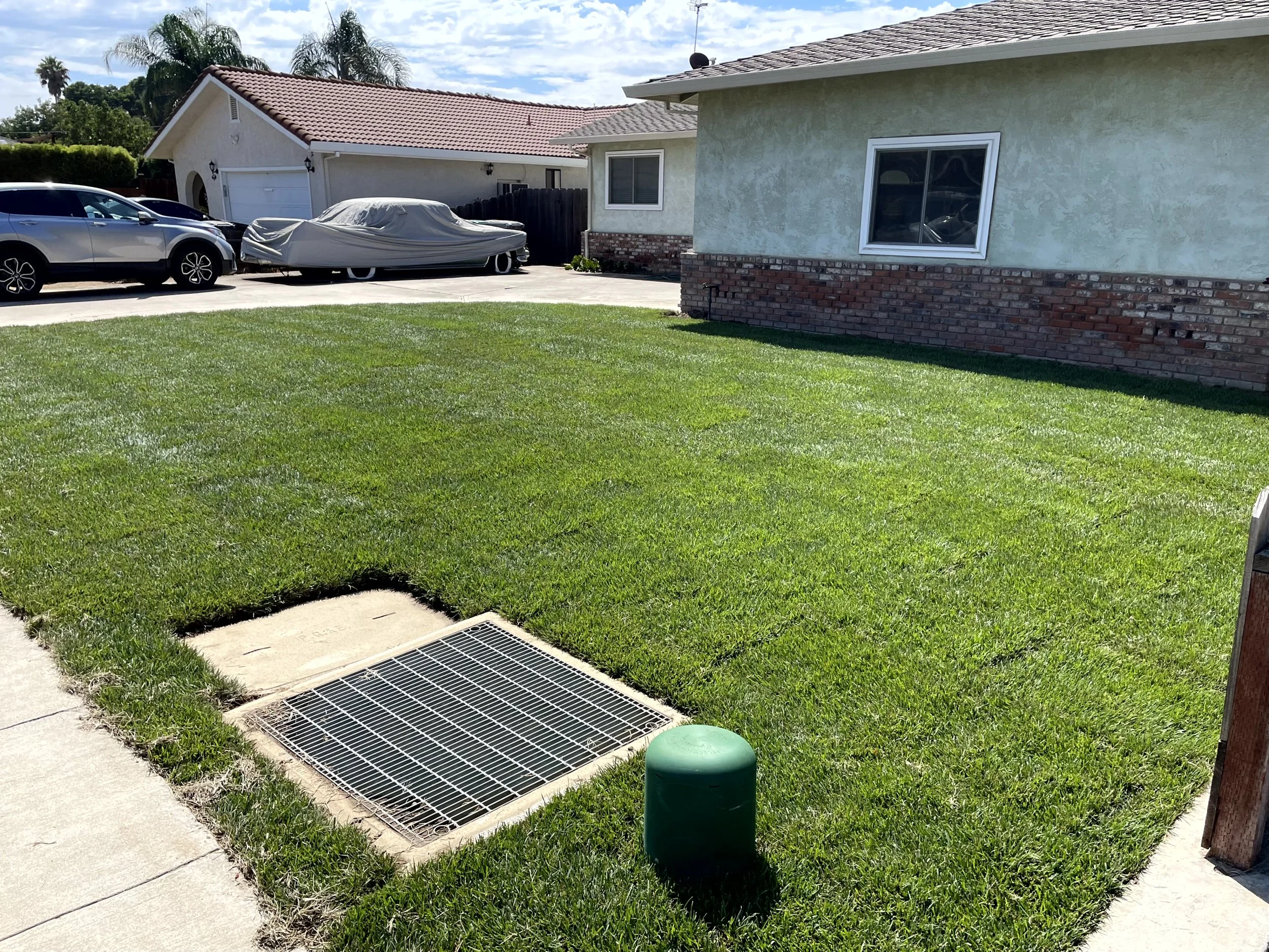 Front yard with green grass, a vent grate, and a green water meter cover, with houses and cars in the background.