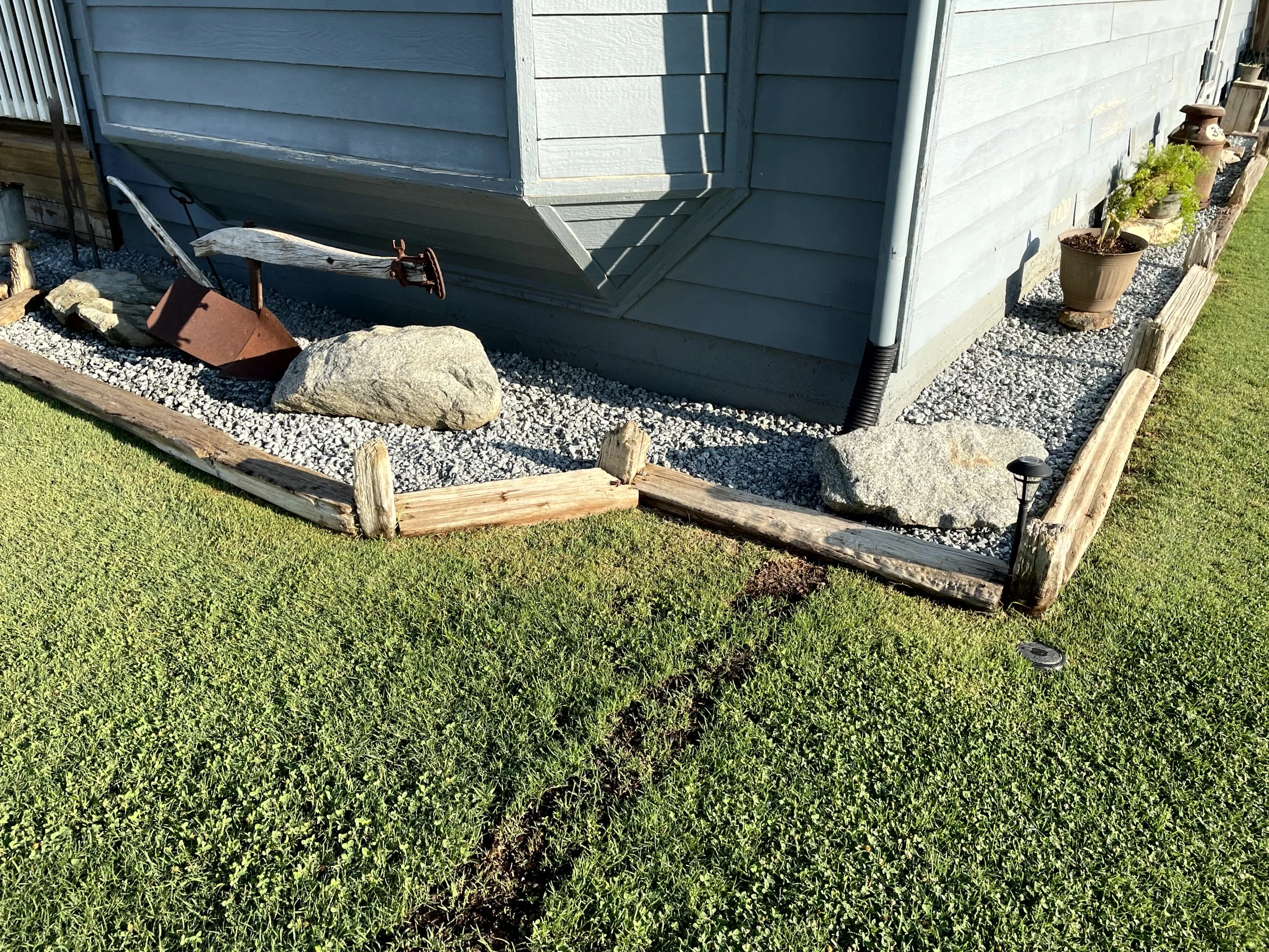 Corner of a house with a landscaped yard, featuring a small garden bed with gravel, large rocks, potted plants, and a weathered garden shovel and rusted watering can.