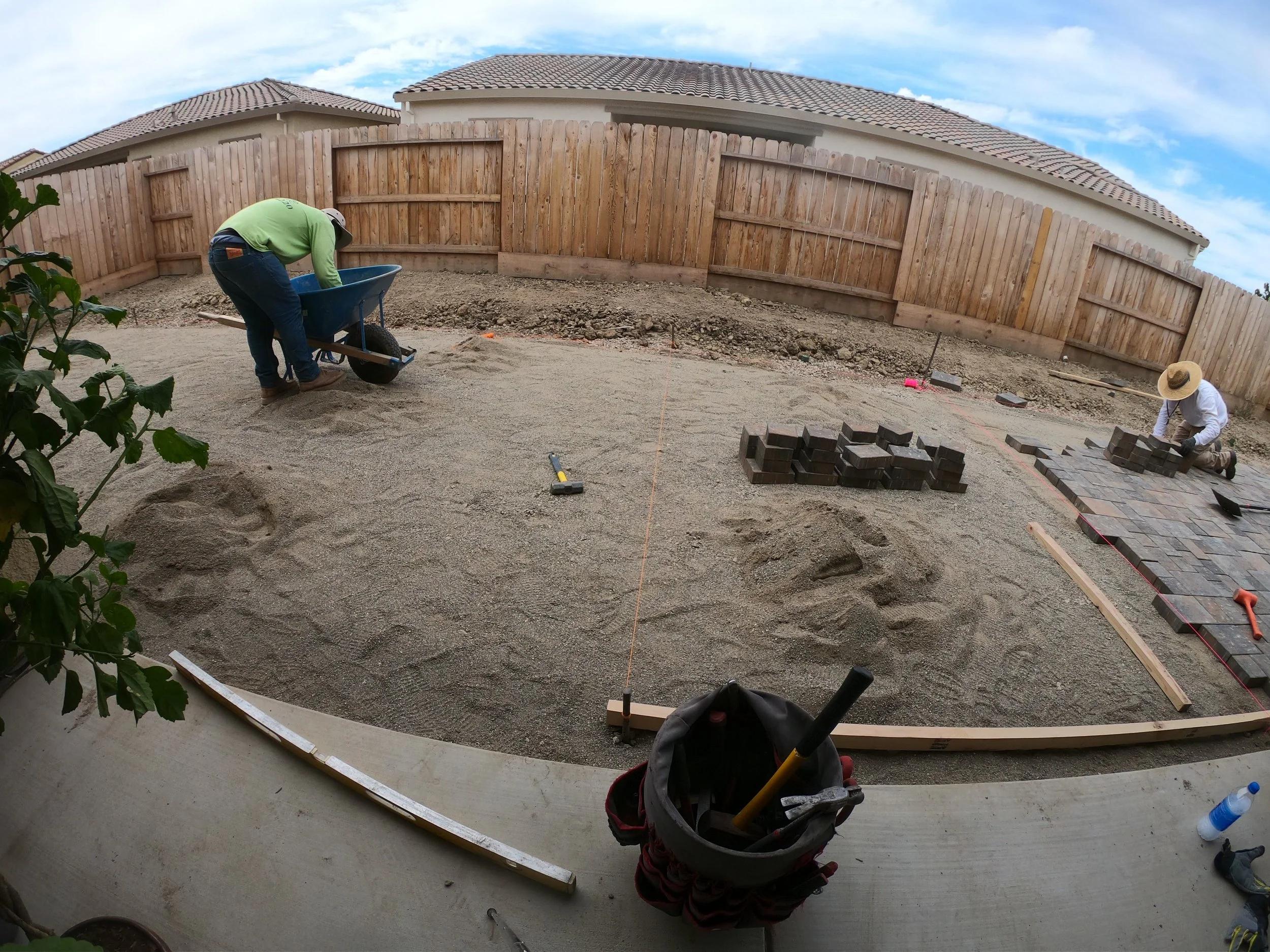 Two workers are constructing a stone patio in a backyard, with one pushing a wheelbarrow and the other carefully laying bricks on the ground.