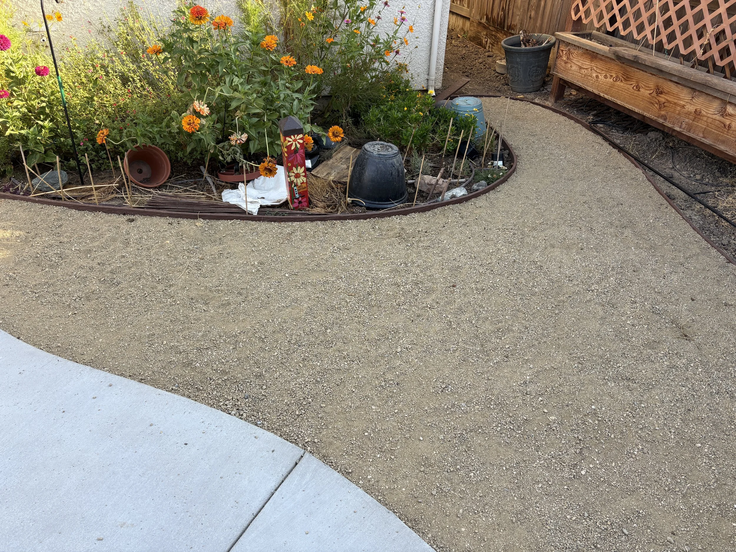 Garden bed with orange and pink flowers, surrounded by a gravel pathway, some pots, and garden tools.