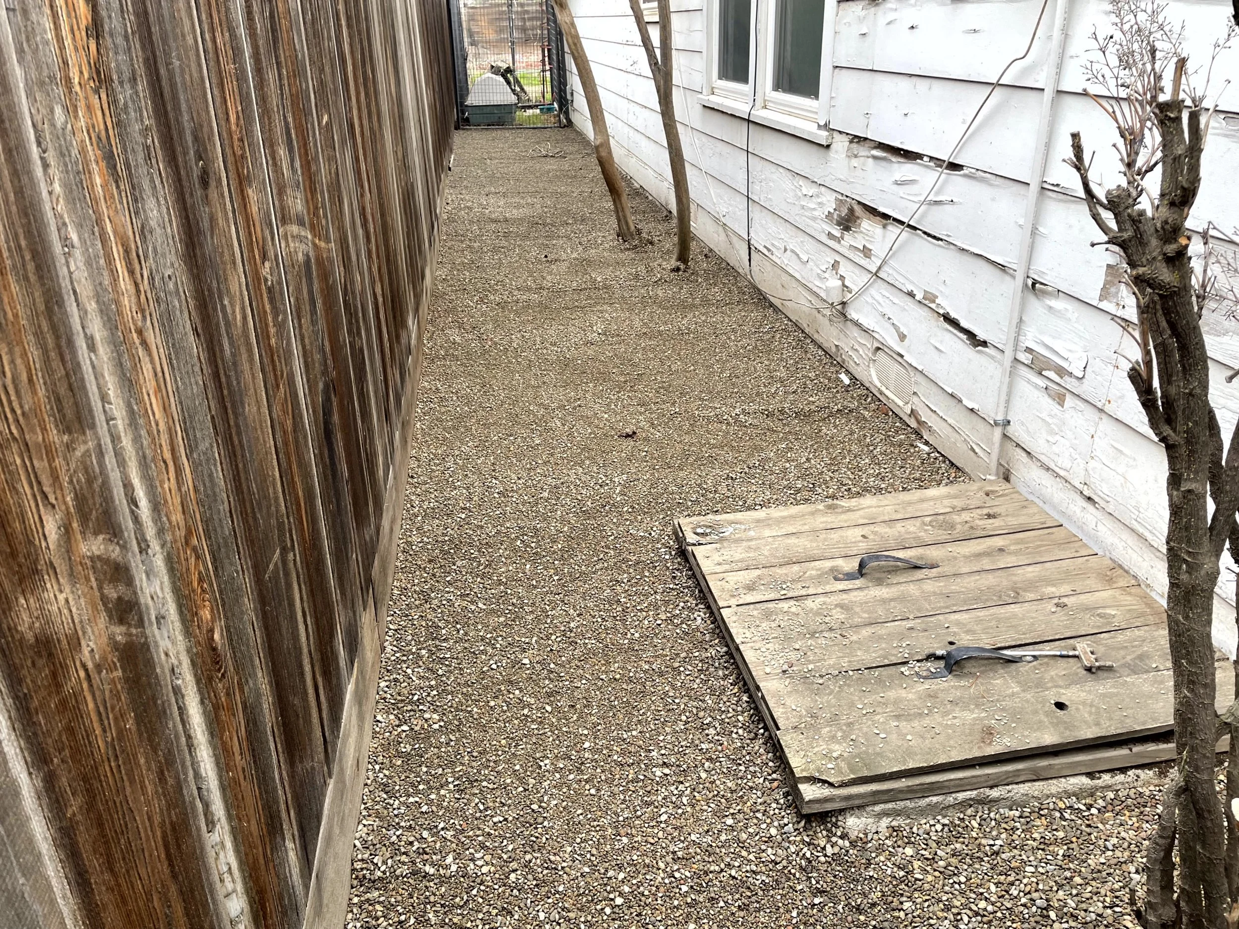 An outdoor backyard alleyway with gravel ground, a wooden fence on the left, a white worn house wall with peeling paint on the right, a small leafless tree on the right, a wooden platform on the ground, and tools resting on the platform. In the backg