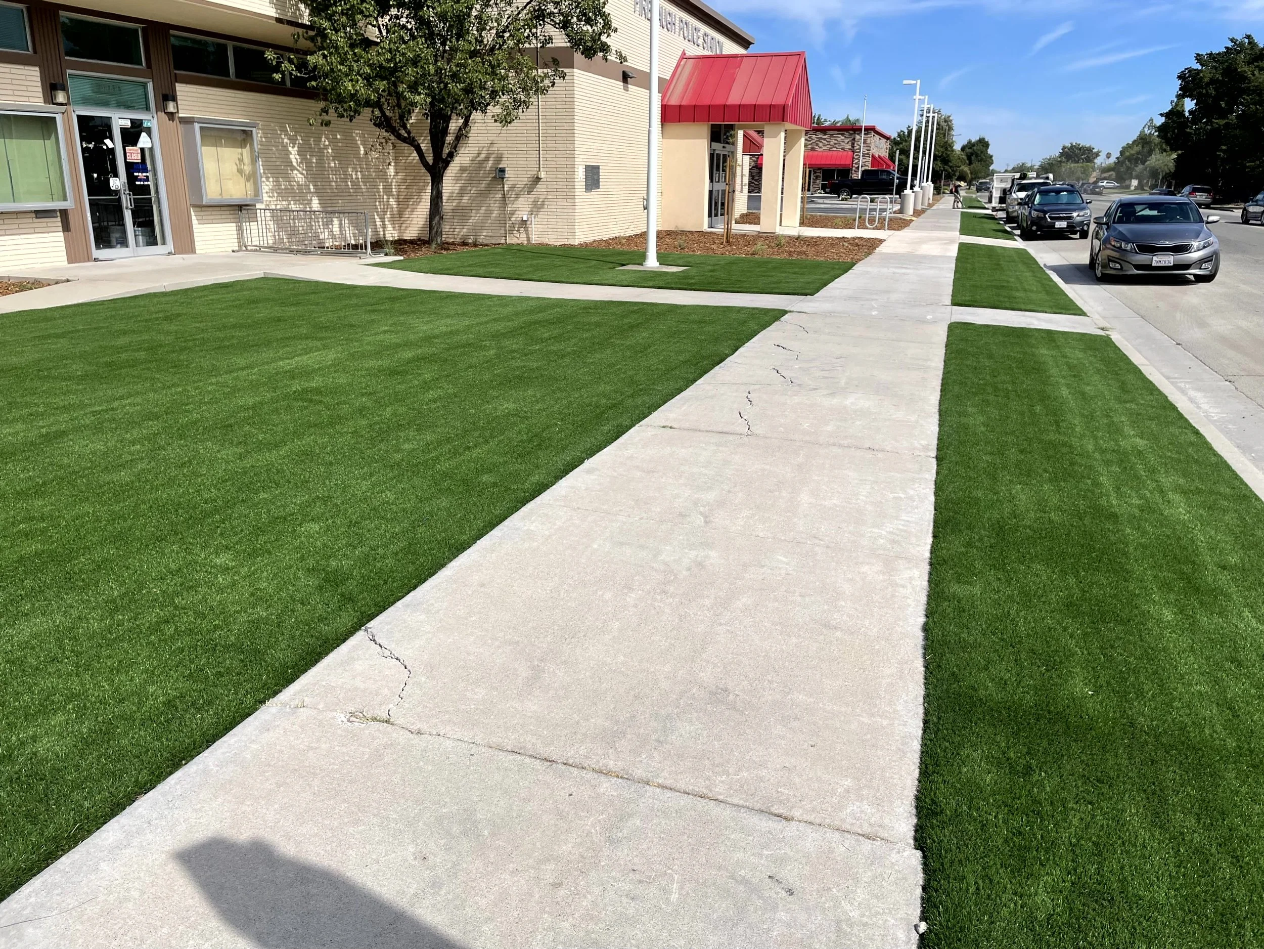 Sidewalk with fresh green grass on either side, in front of a building with a red roof and parked cars.