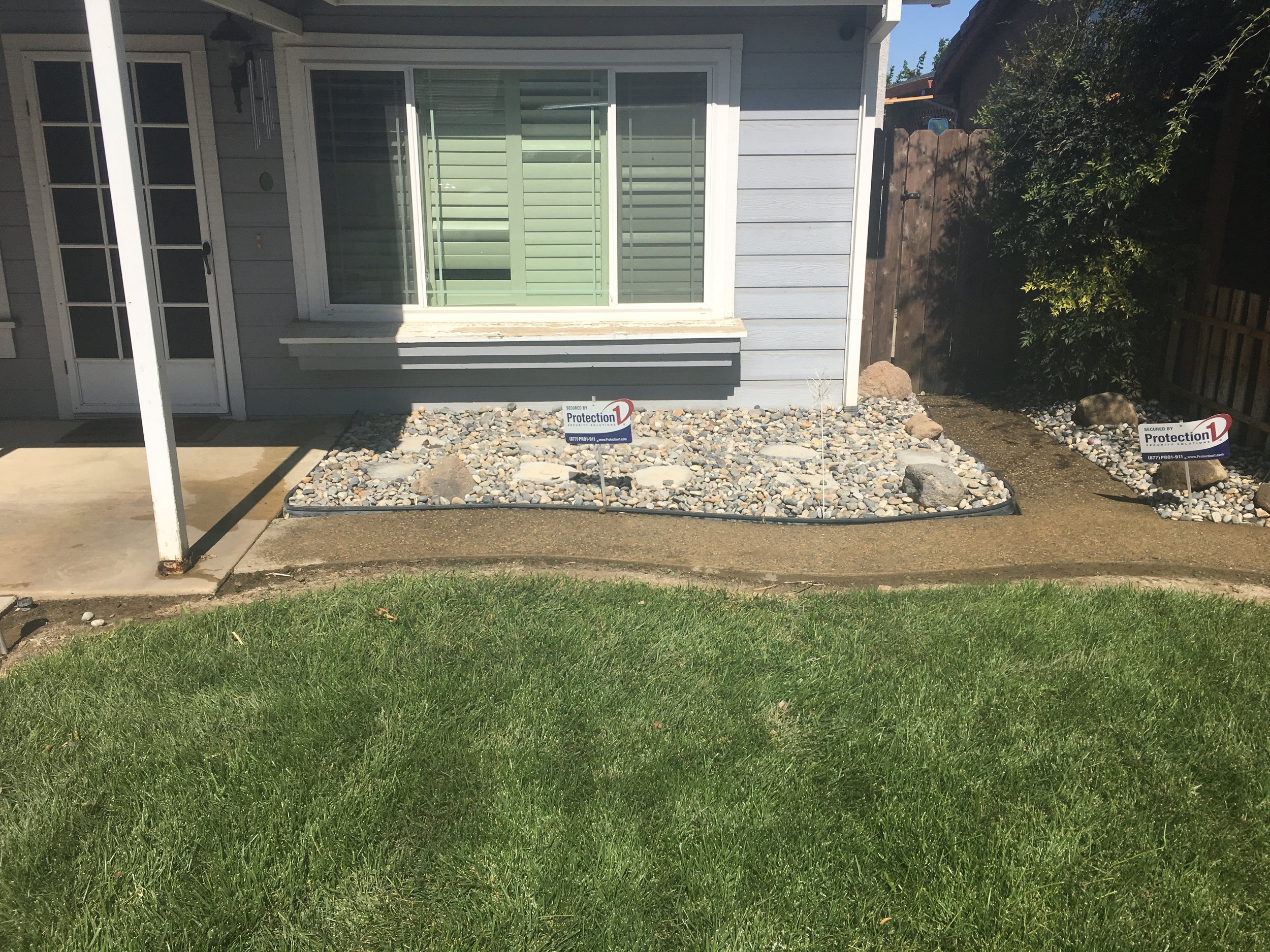 Front yard landscaping with new rock beds, bordered by a black plastic edging, and placed in front of a house with a window and door, with signs indicating security by Protection 1.