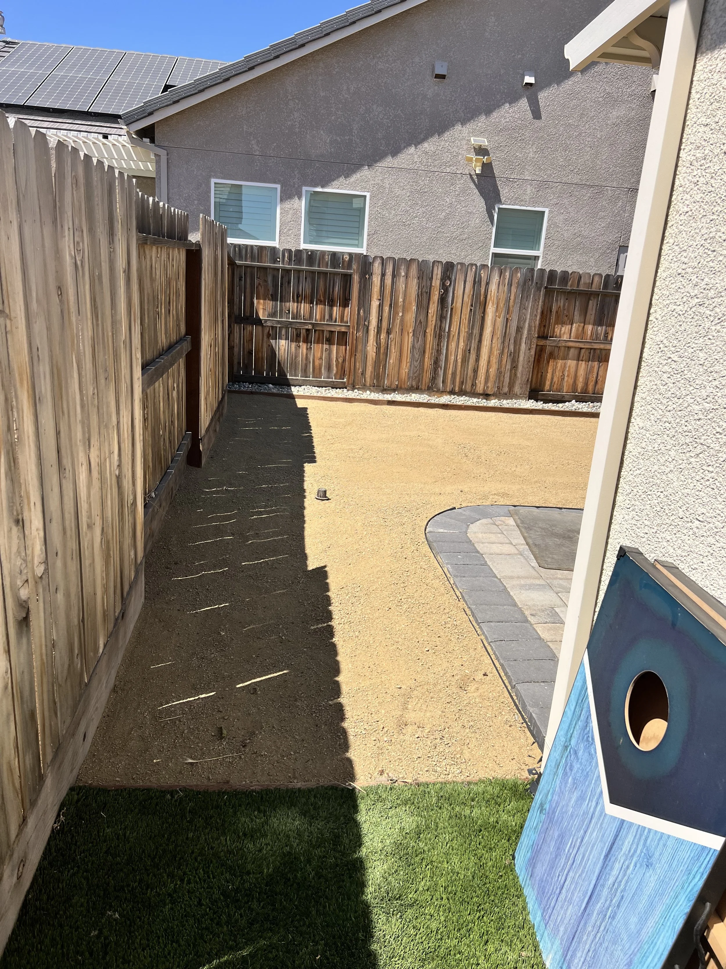 A backyard with a fenced area, dirt ground, a small patch of artificial grass, and a partially completed cornhole game set. The background shows a house with solar panels on the roof and two small windows.