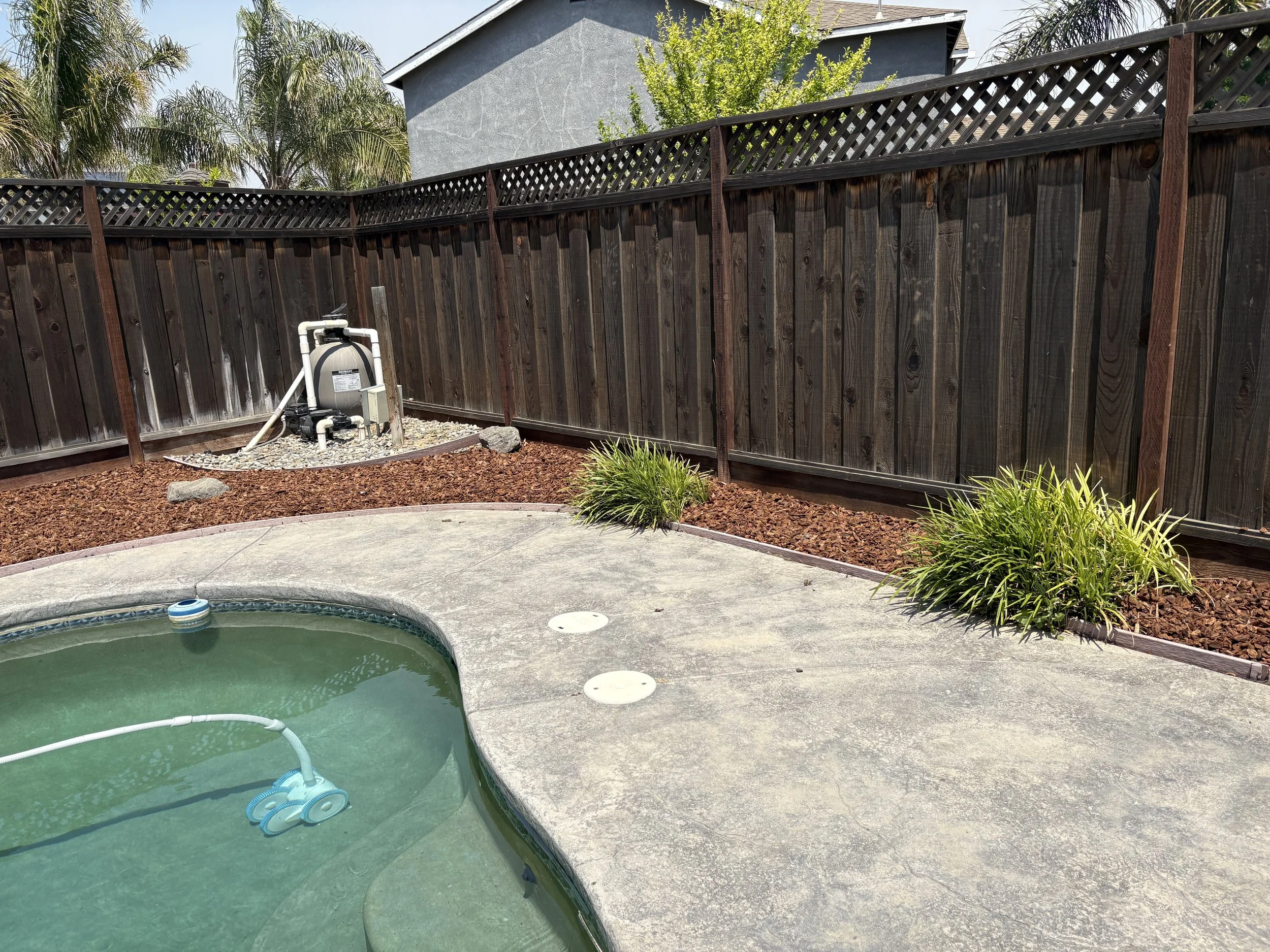 Backyard with a small, empty pool, a wooden privacy fence, some plants, and a pool filtration system in a corner.