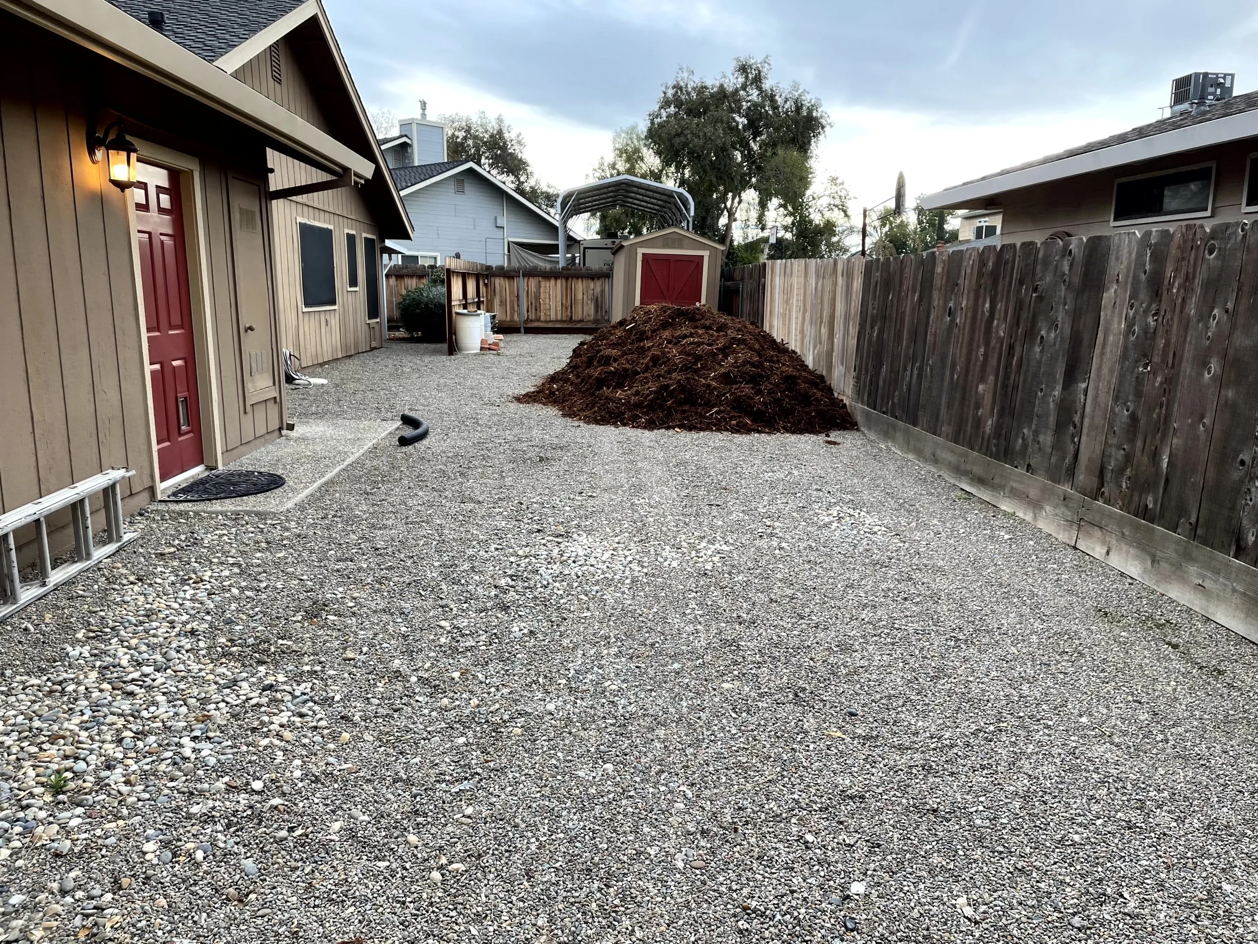Backyard with gravel ground, a red door on a beige house, a fenced yard, and a large pile of mulch or soil near the fence. There are some garden tools, a ladder, and a shed in the background.