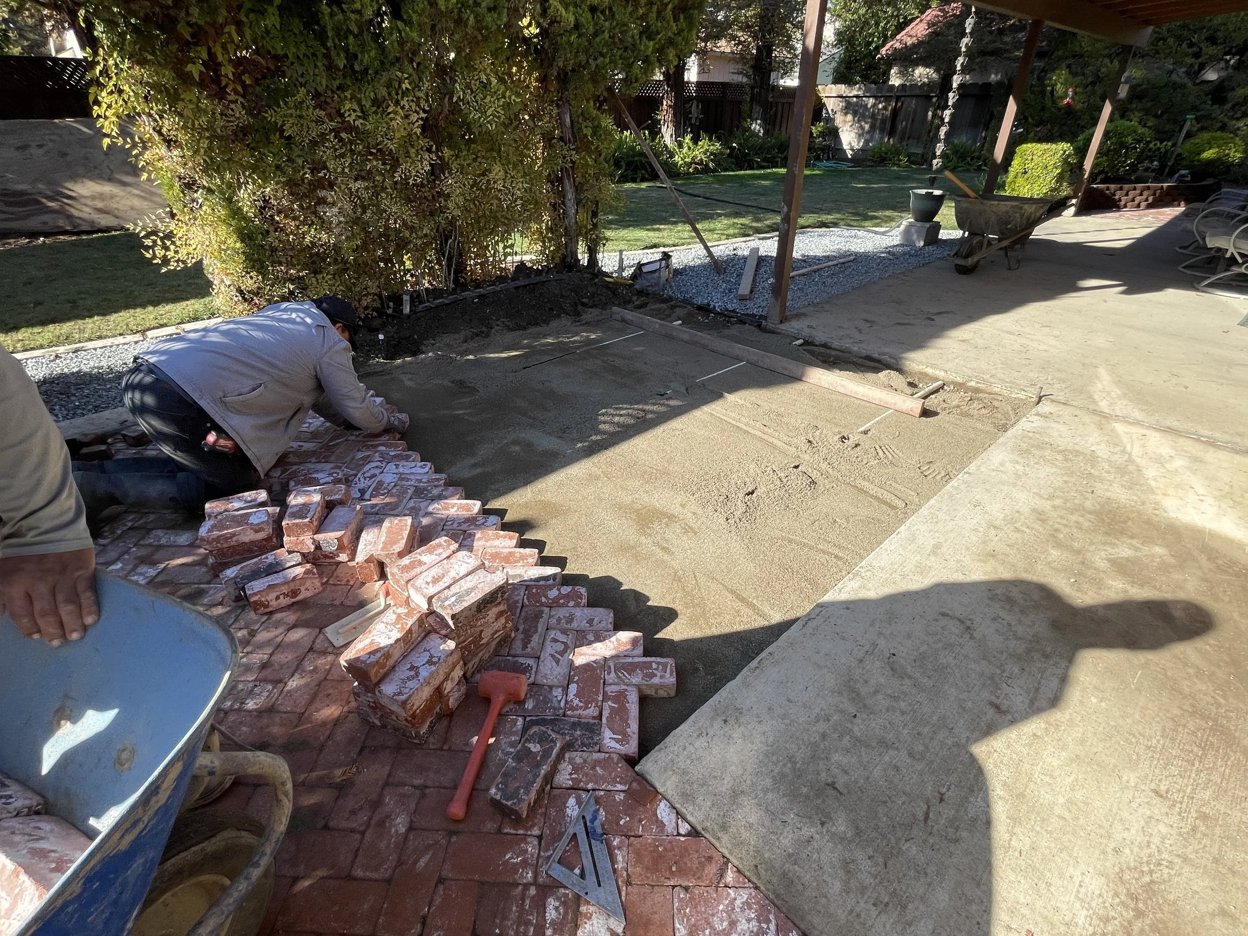 Person laying bricks on a garden patio, preparing to lay concrete or pavers, with construction tools and materials visible nearby, in a backyard setting with trees and a fence.