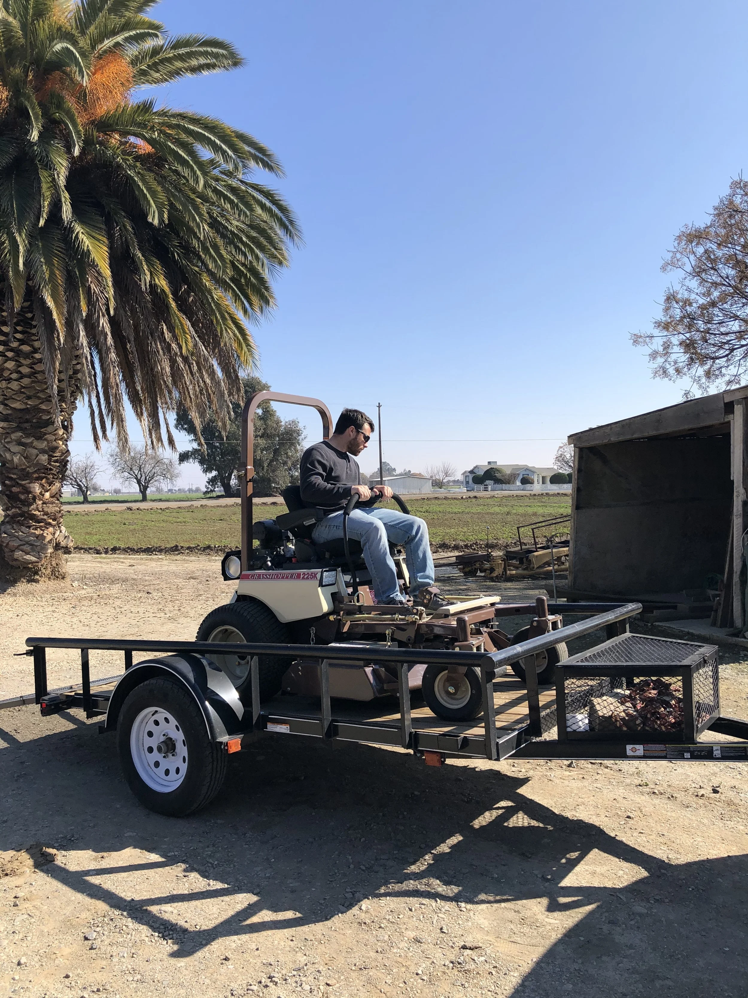 A person driving a lawn mower on a trailer in a rural area with palm trees and a barn in the background.