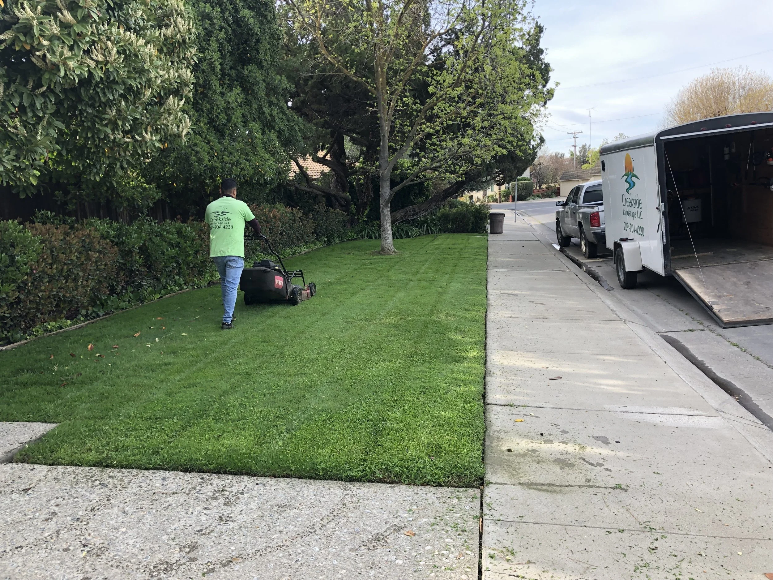 A worker in a bright green T-shirt is mowing a lush, well-manicured lawn next to a sidewalk. There are large green bushes and trees in the background, and a white moving truck with a ramp parked on the street.