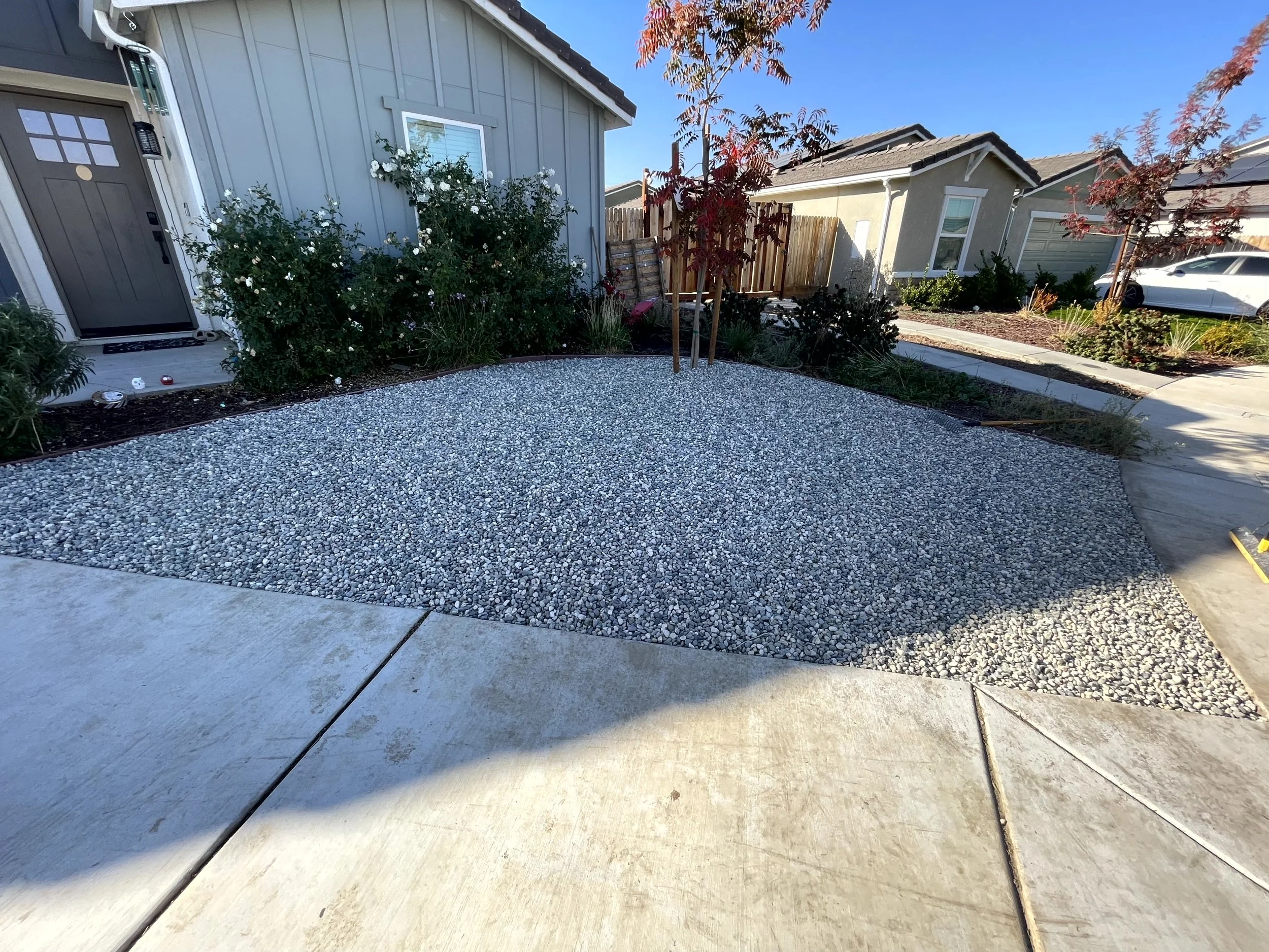 A front yard with a gravel area surrounded by concrete and a house in the background. There are small trees and bushes along the house and a wooden fence.