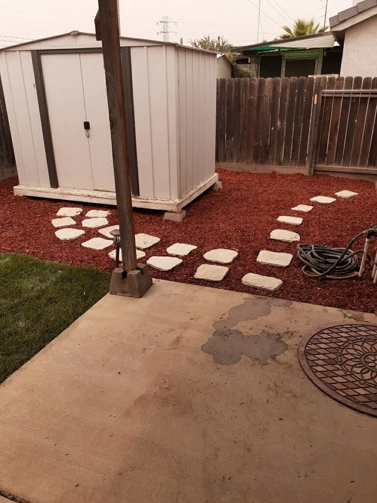 Backyard with concrete patio, stepping stone path leading to a storage shed, garden hose coiled on the ground, wooden fence, and adjacent lawn area.