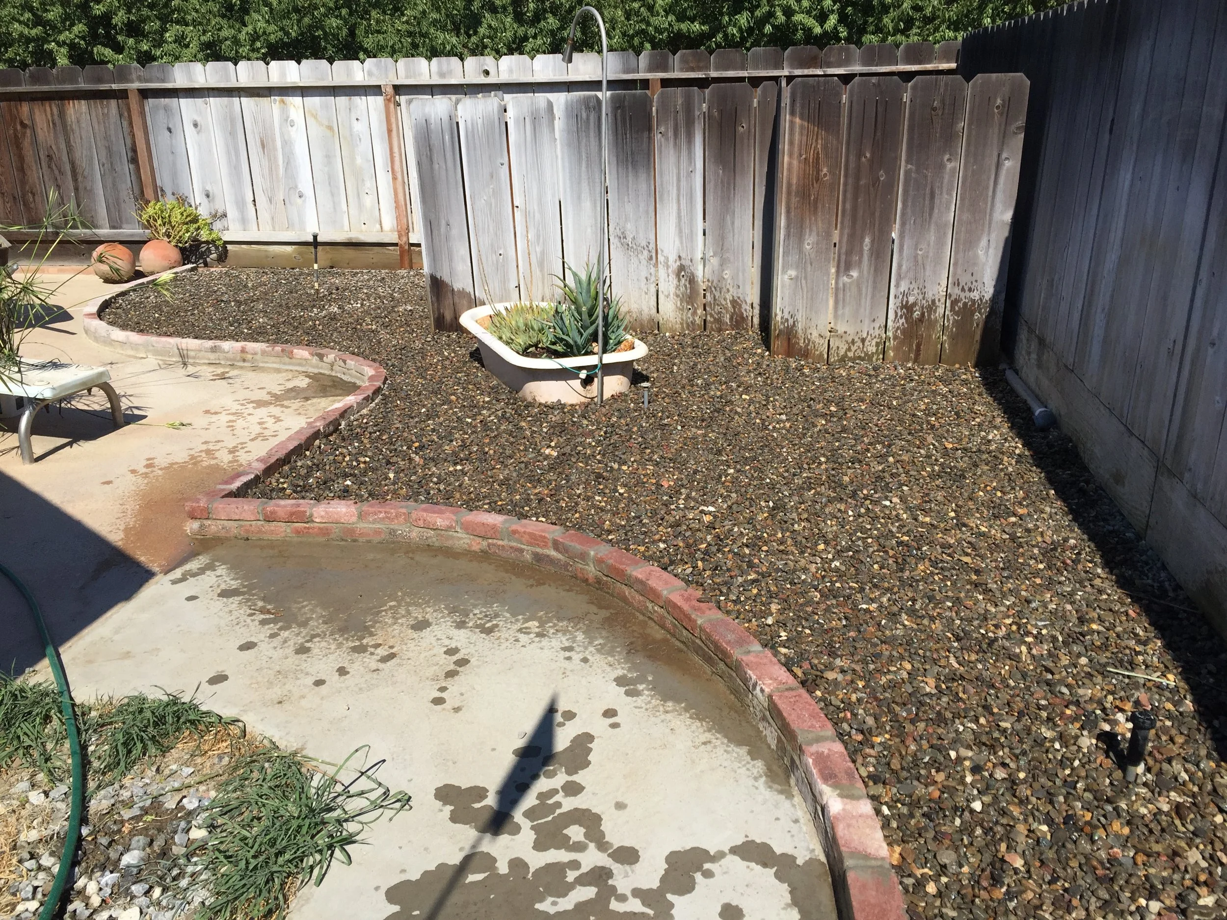 Backyard garden with a gravel bed, a plant in a white tub, a concrete patio with a curved brick border, wooden fence, and a small solar light.