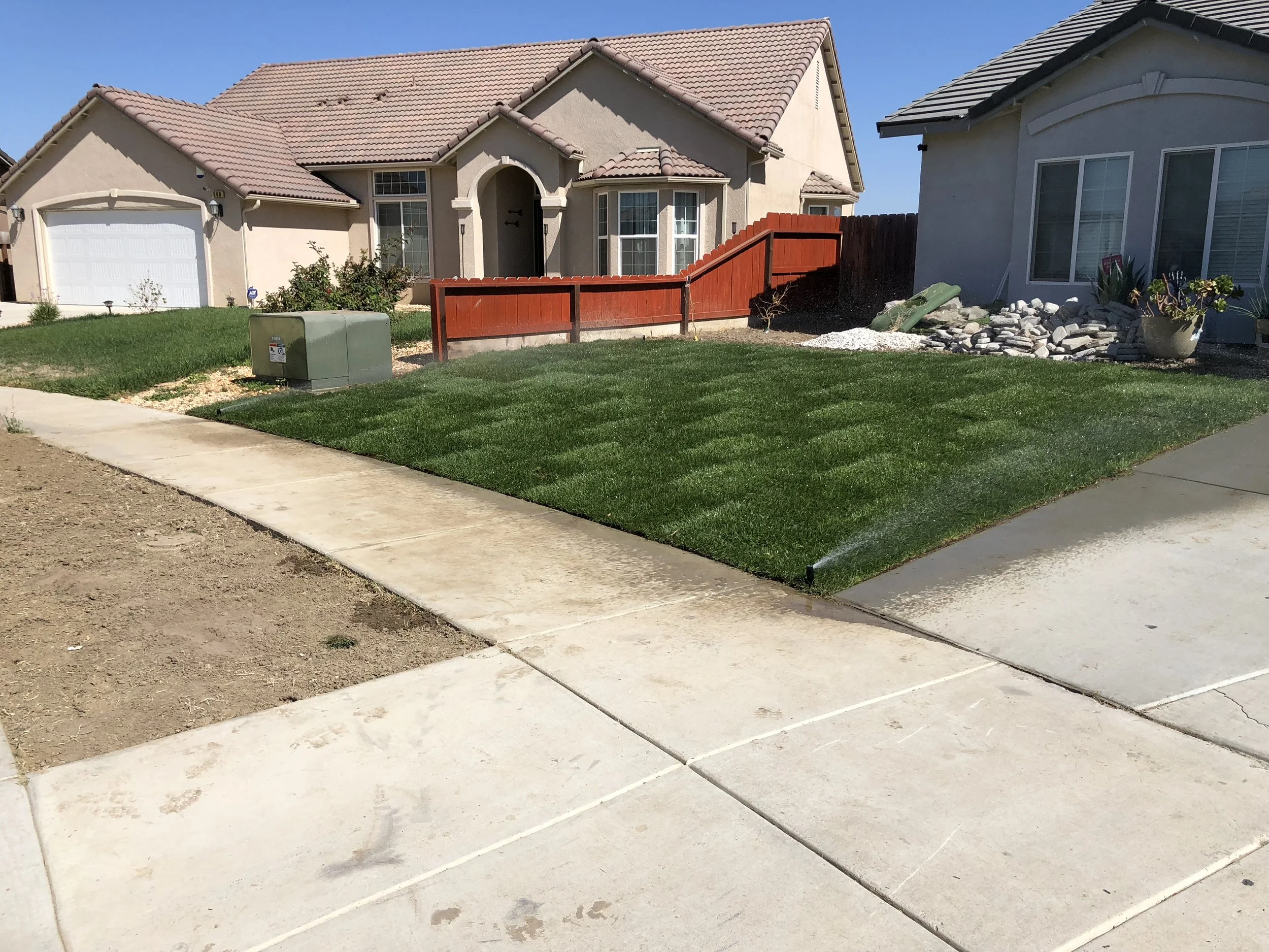 A residential front yard with a lush green lawn, a concrete sidewalk, and a house with a tiled roof. There is a small garden area with rocks and plants on the right side.